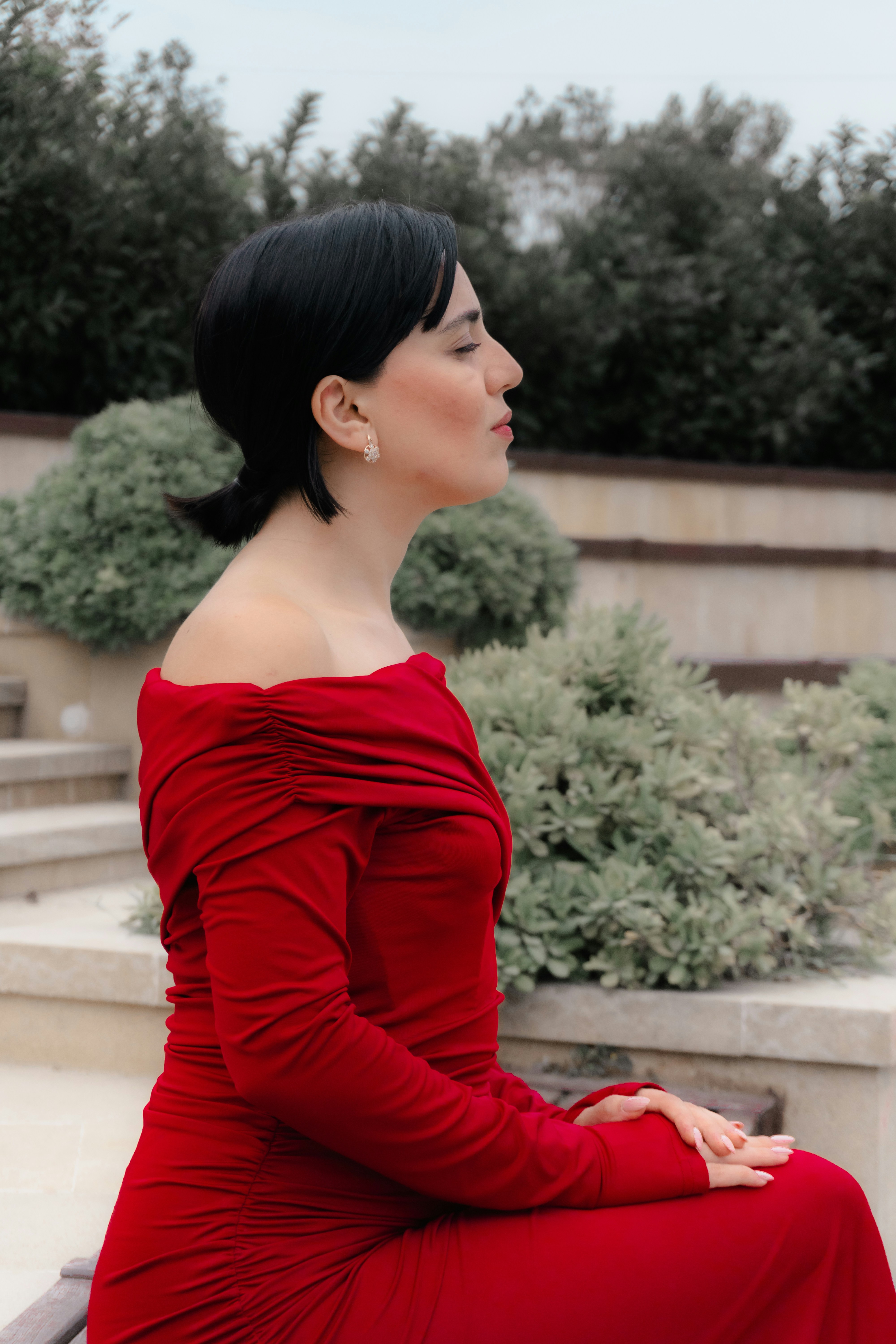 A woman in a red off-the-shoulder dress sitting outdoors with eyes closed