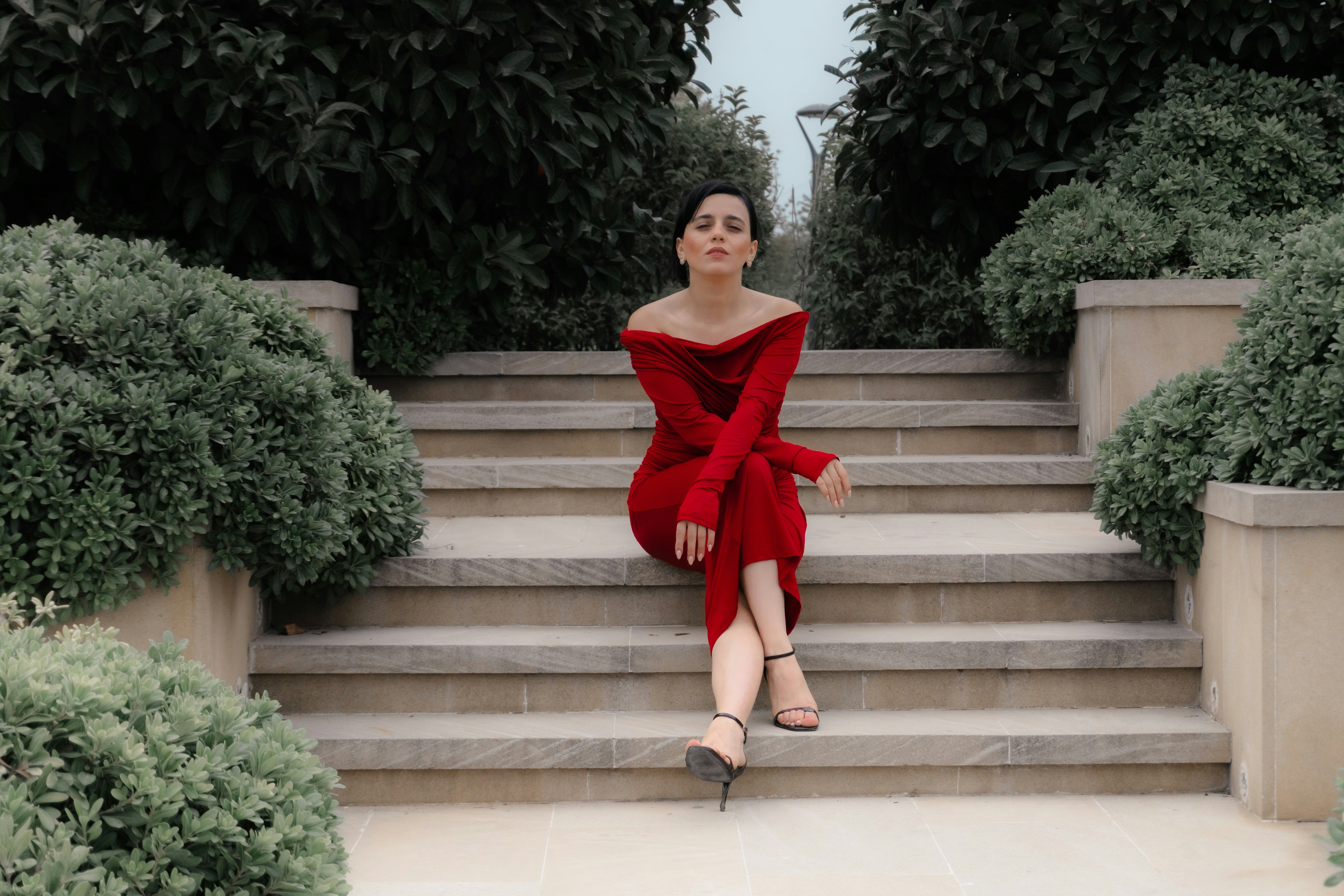 A woman in a red off-the-shoulder dress sitting on stone steps between bushes