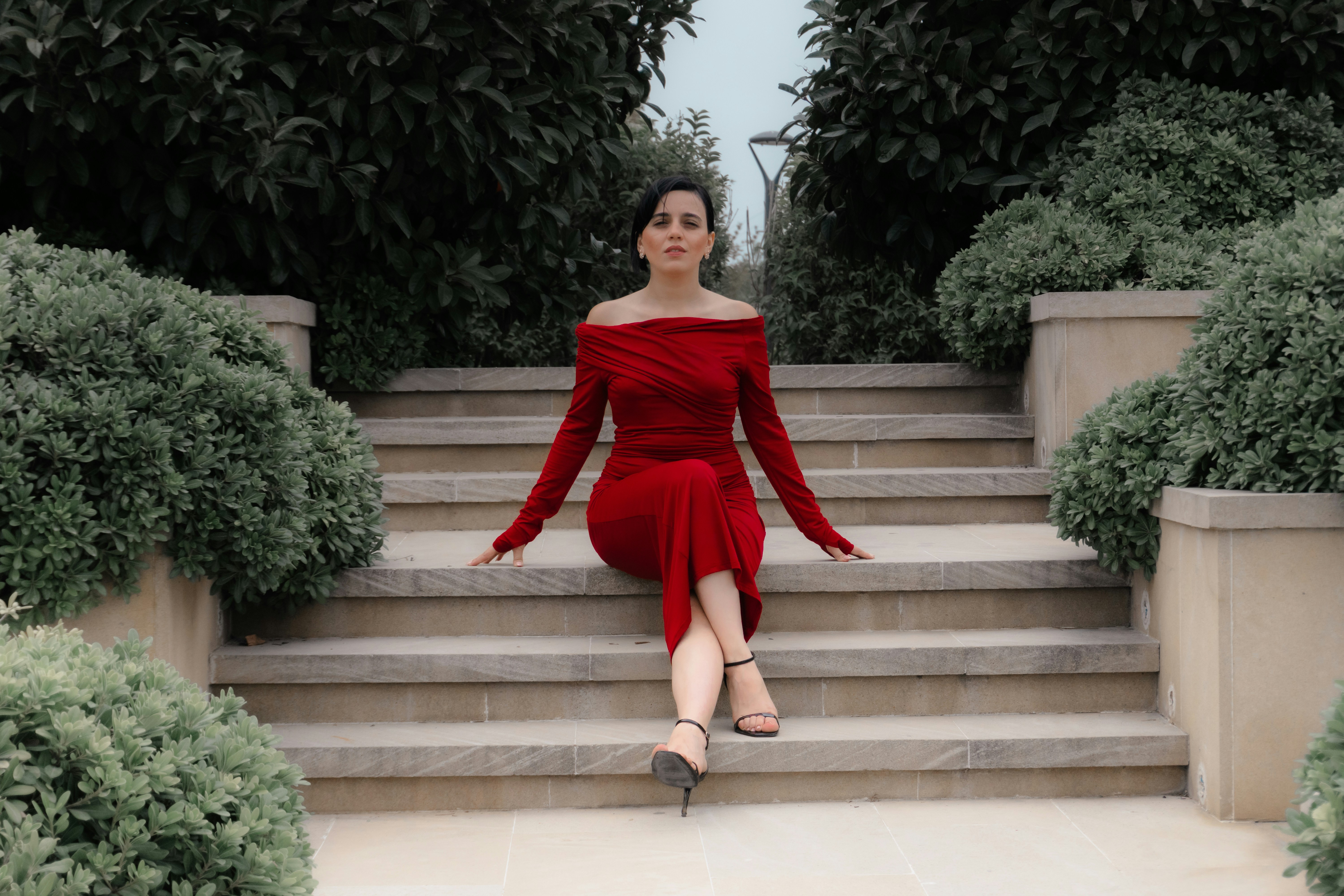 A woman in a red off-the-shoulder dress sitting on stone steps between bushes