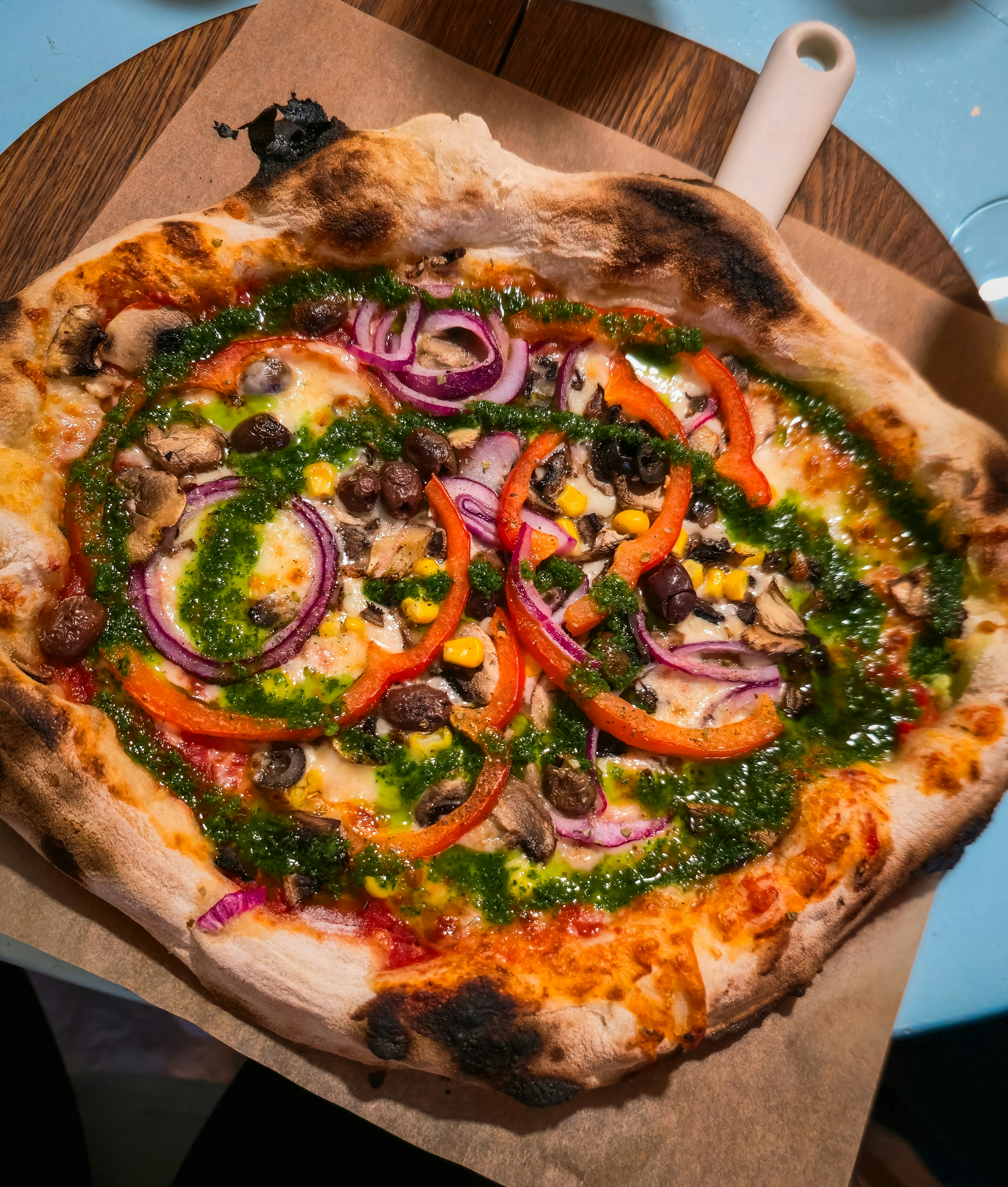 A pizza topped with red peppers, red onions, mushrooms, corn, olives, and green pesto