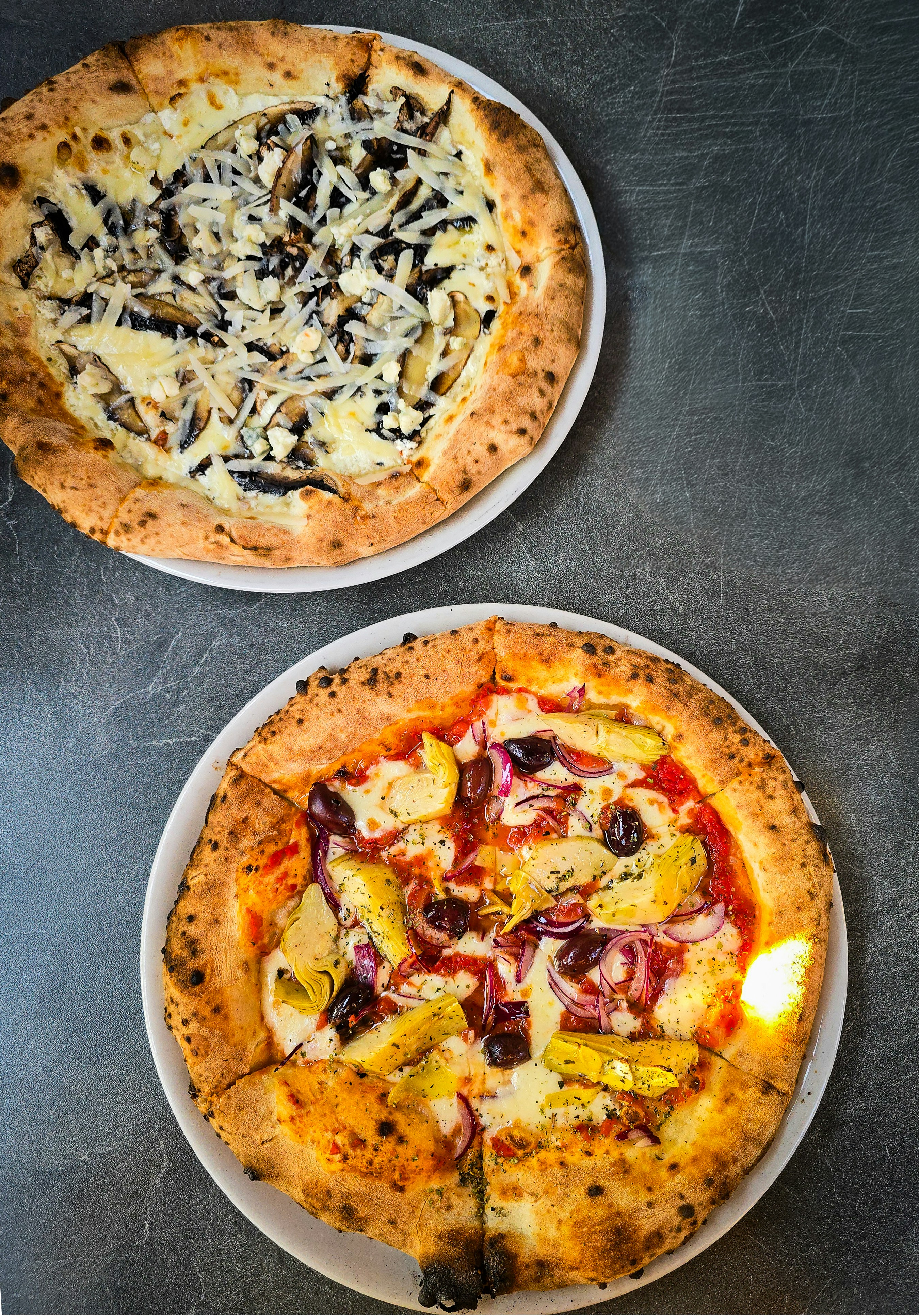 Two pizzas with mushrooms and artichokes on white plates on a dark surface