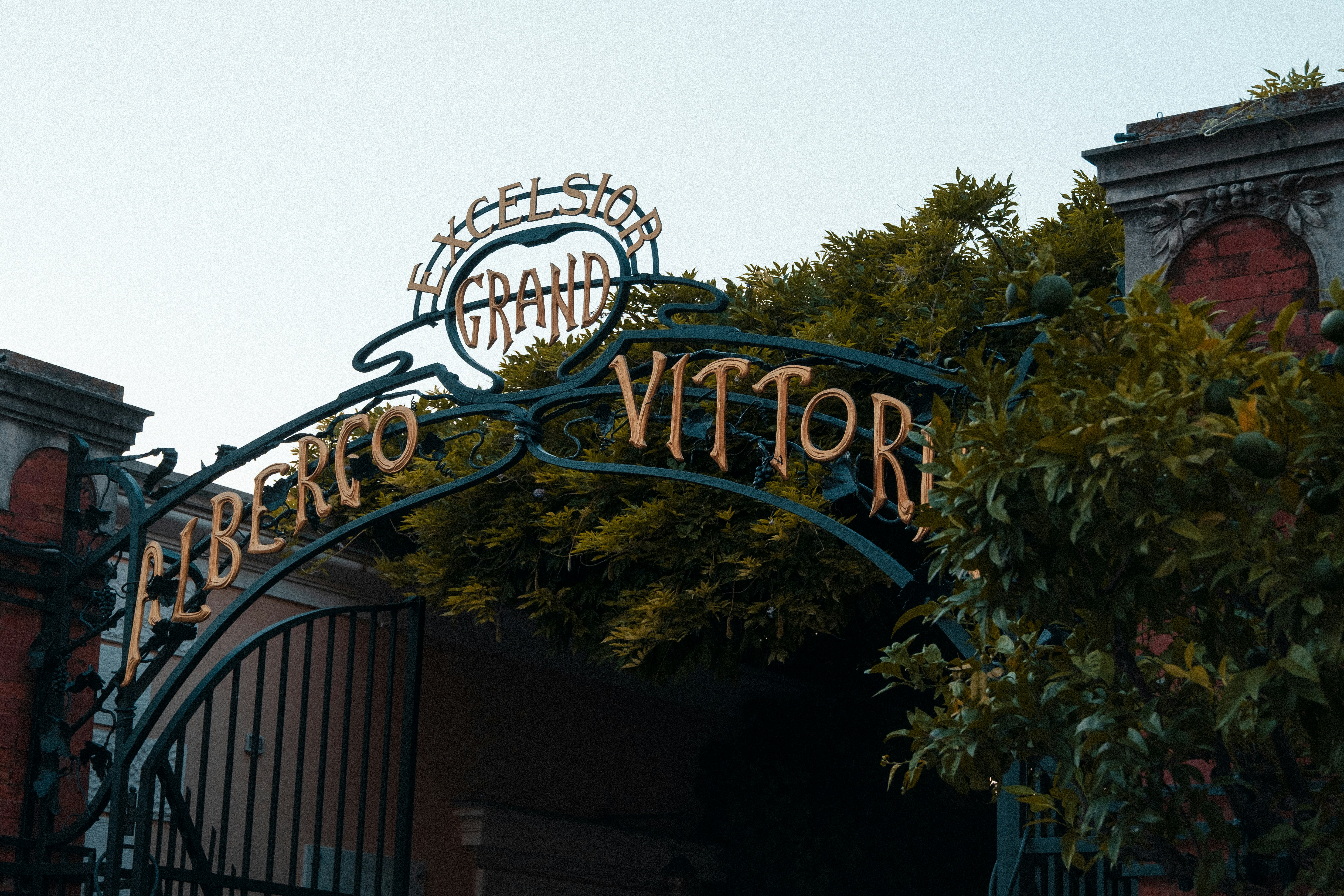 An ornate metal gate reading "Albergo Grand Excelsior Vittoria" beneath lush green foliage