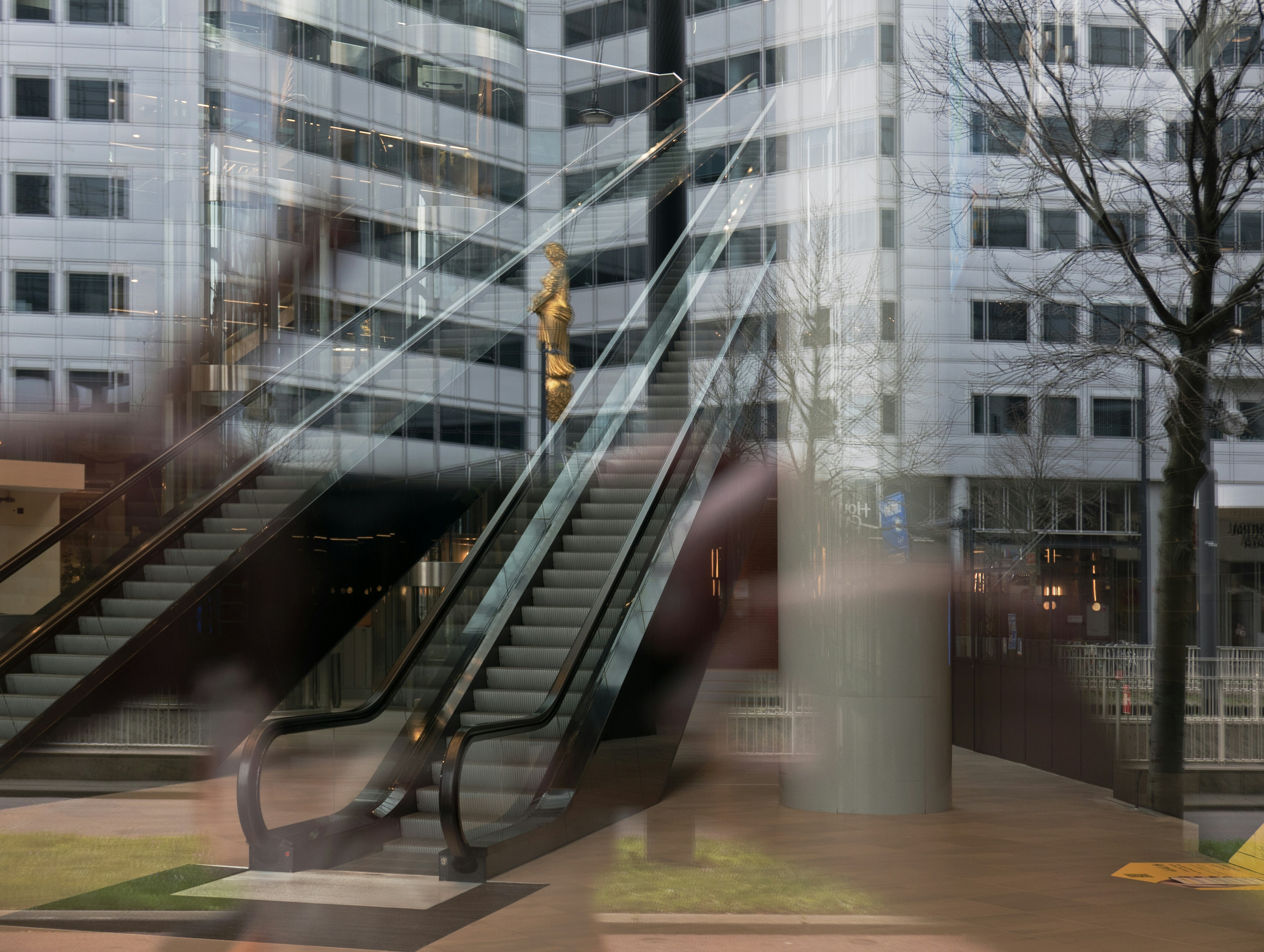 An escalator reflecting in a glass window with a golden statue and office building