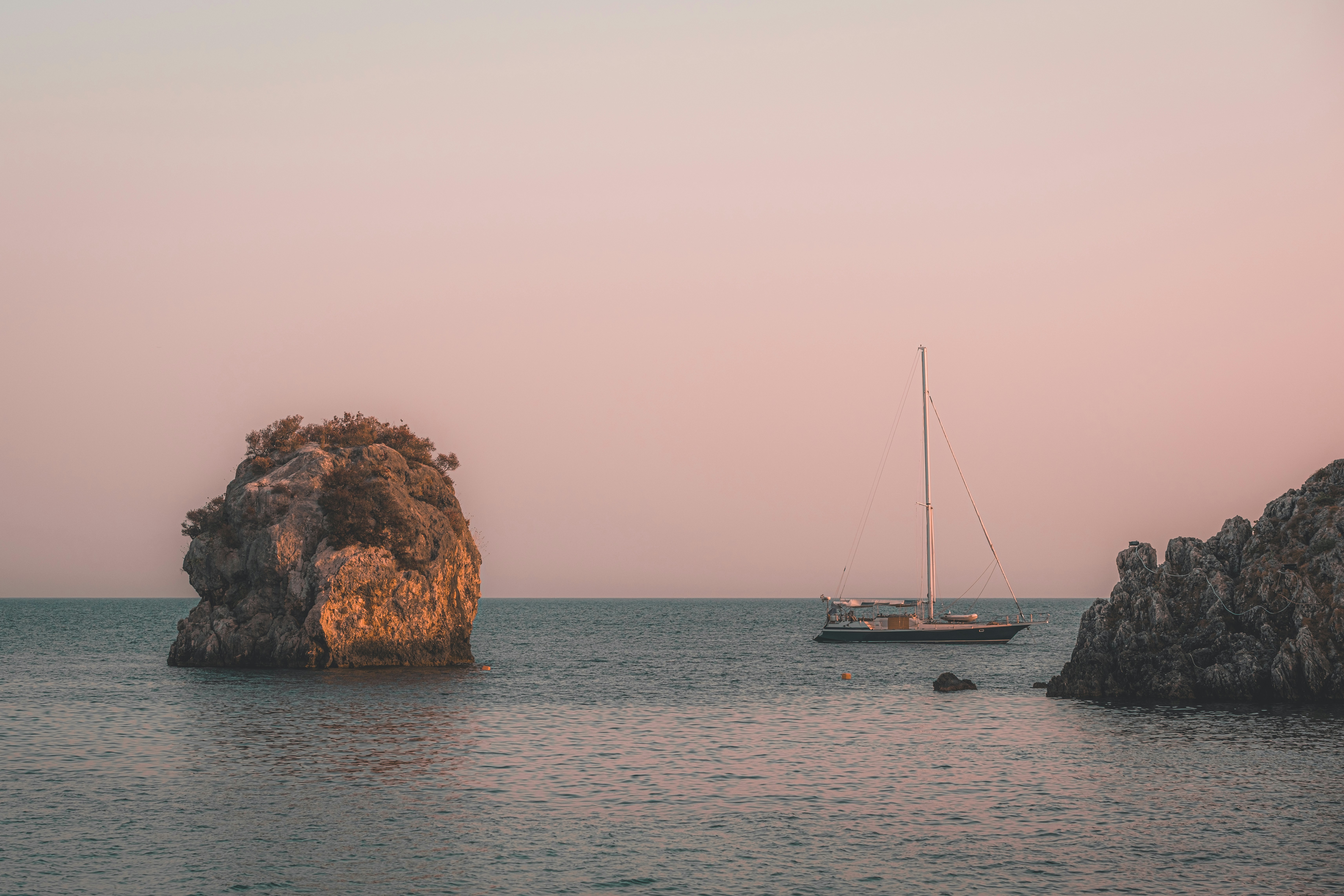 A sailboat on calm water between two rocky outcrops under a soft pink sky