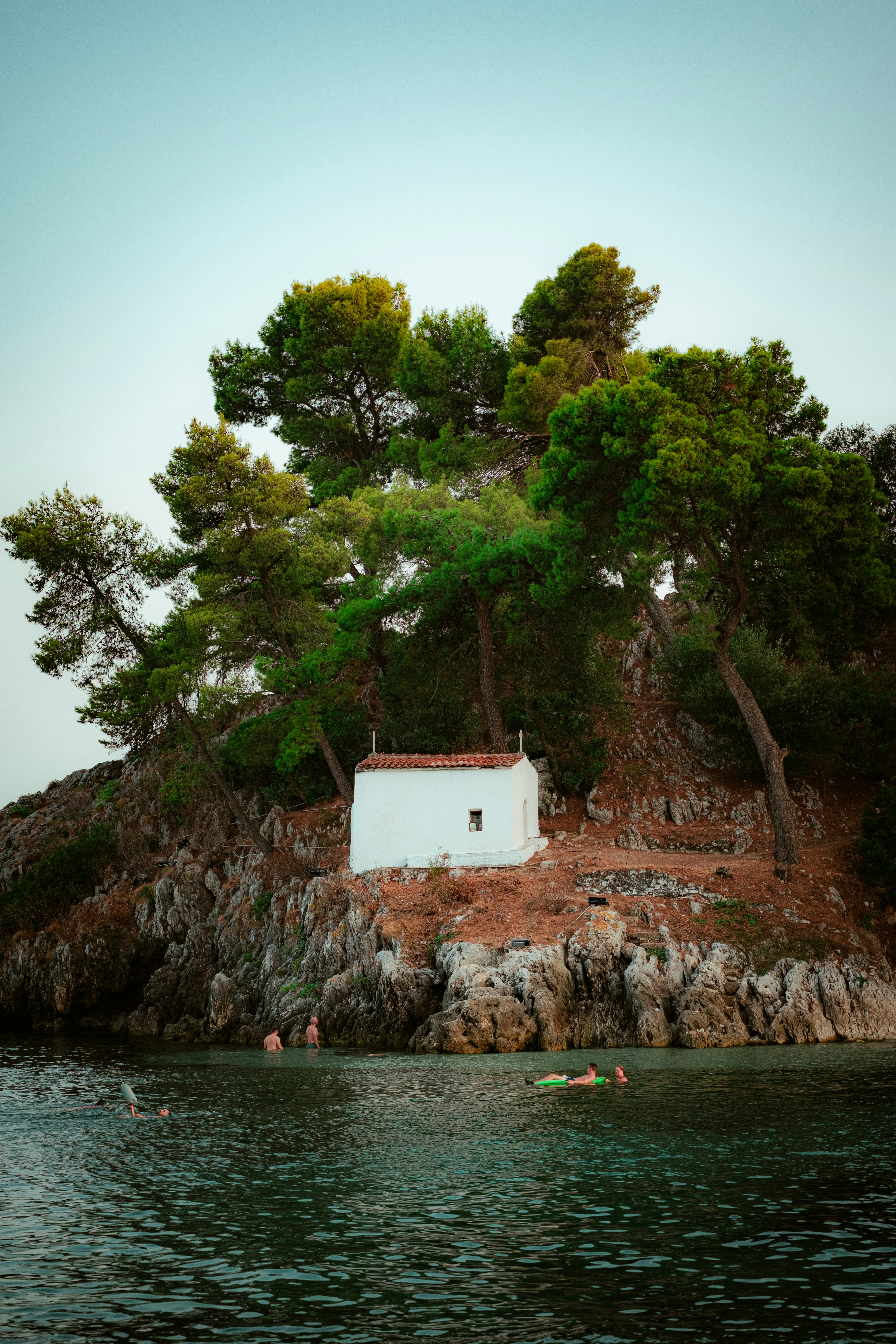 A small white chapel on a rocky cliffside with pine trees above water