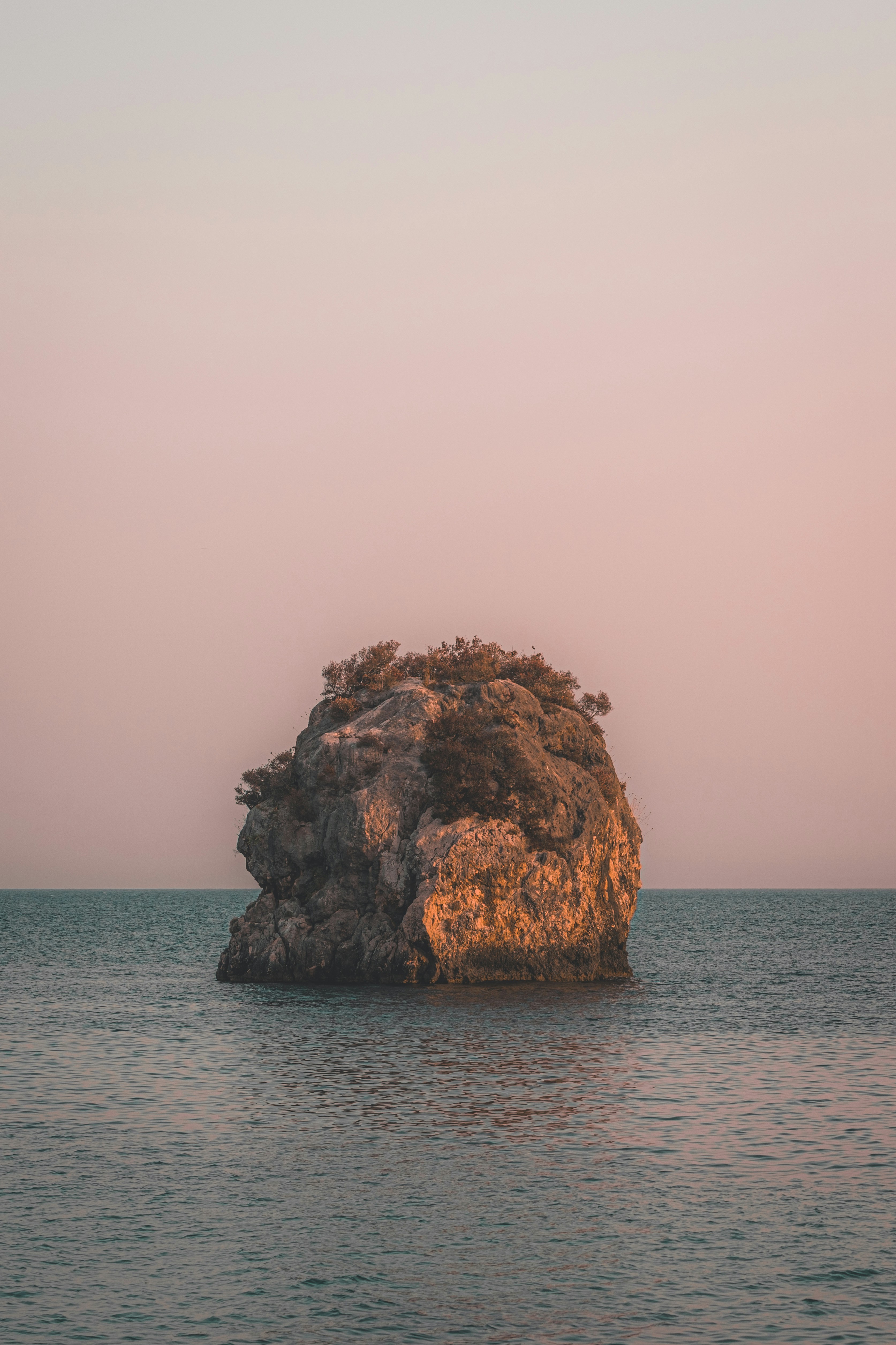 A rocky islet with sparse vegetation in calm water under a pale sky