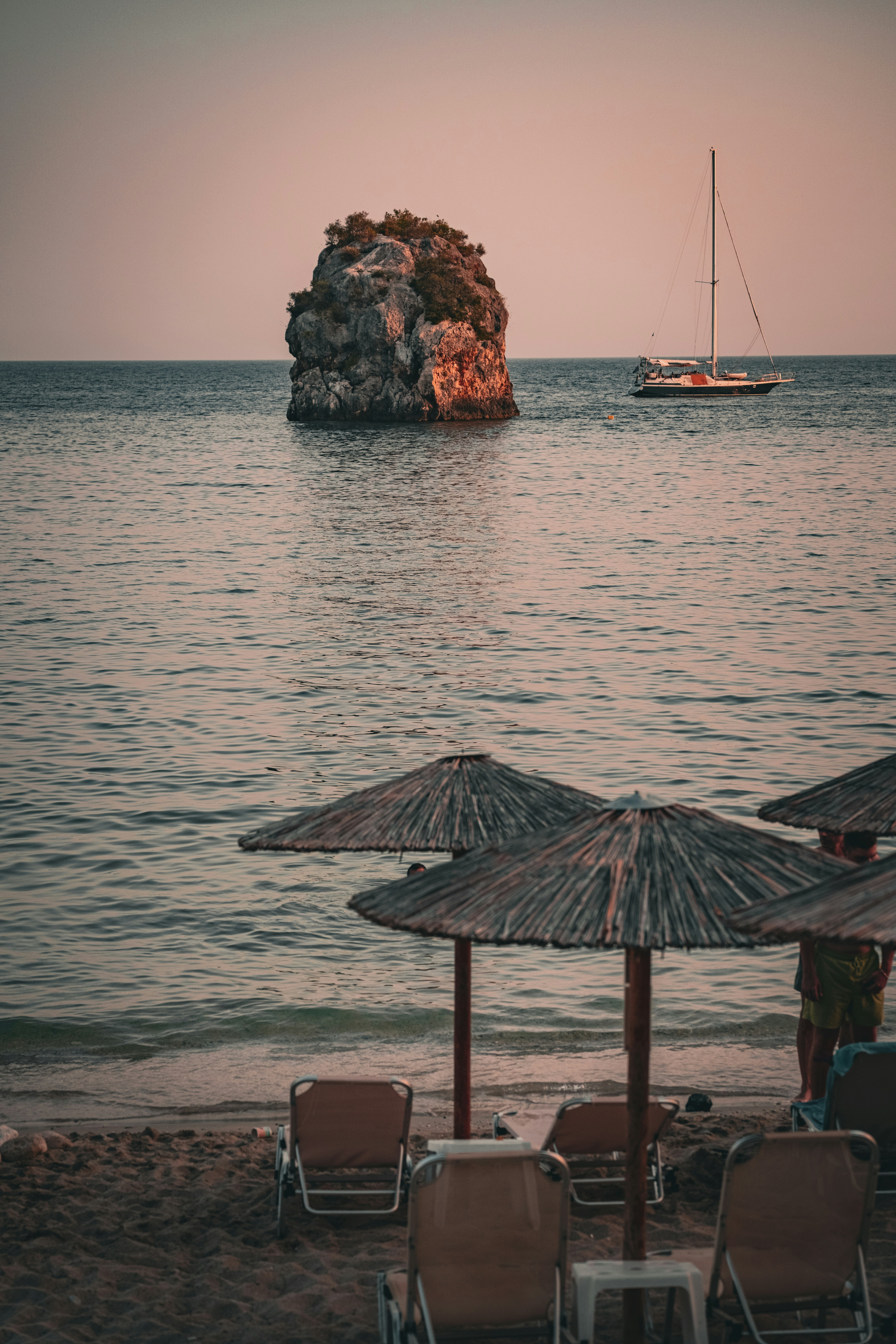 A sailboat near a rocky island viewed from a beach with straw umbrellas