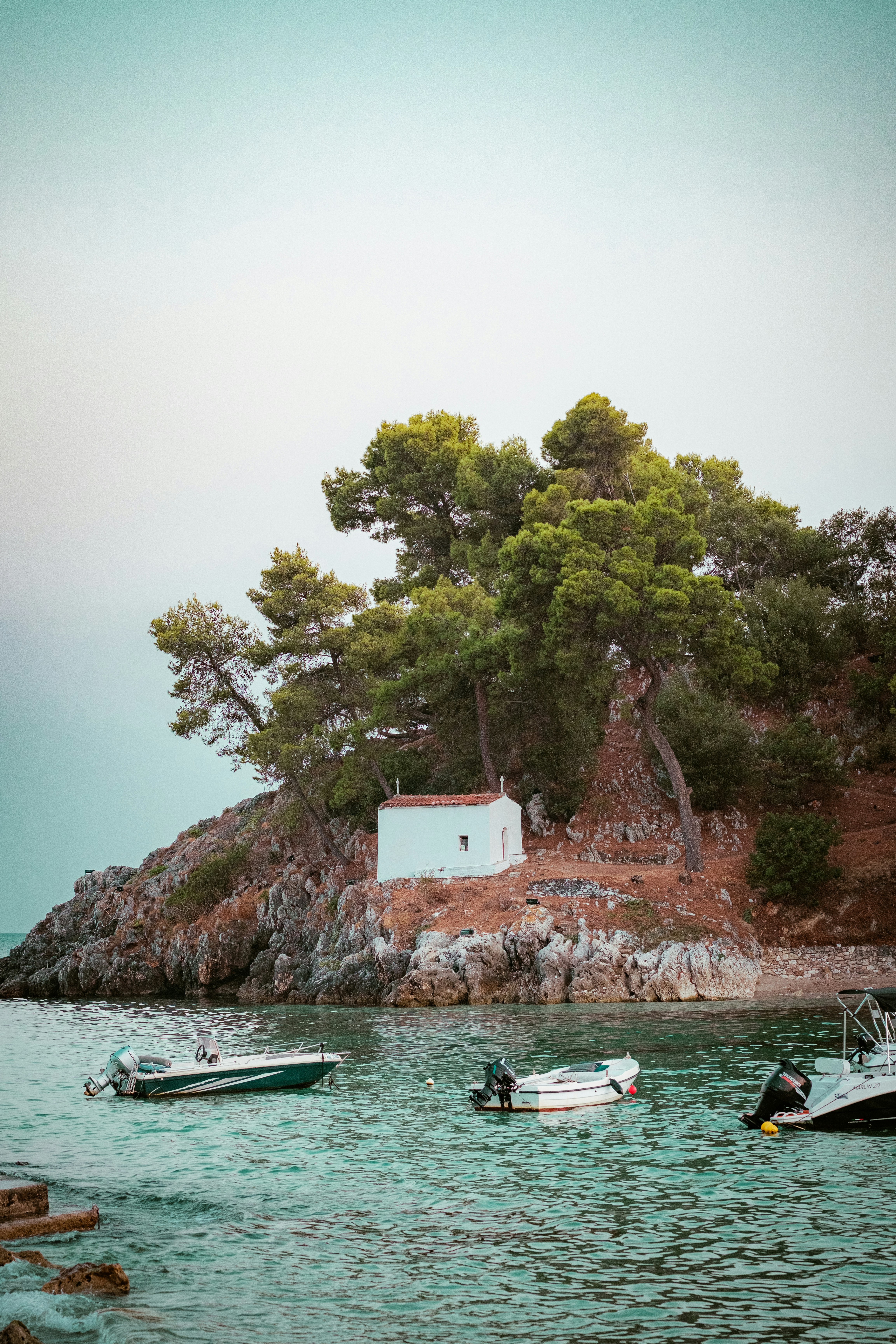 A small white chapel on a rocky pine-covered cliff above turquoise water with boats