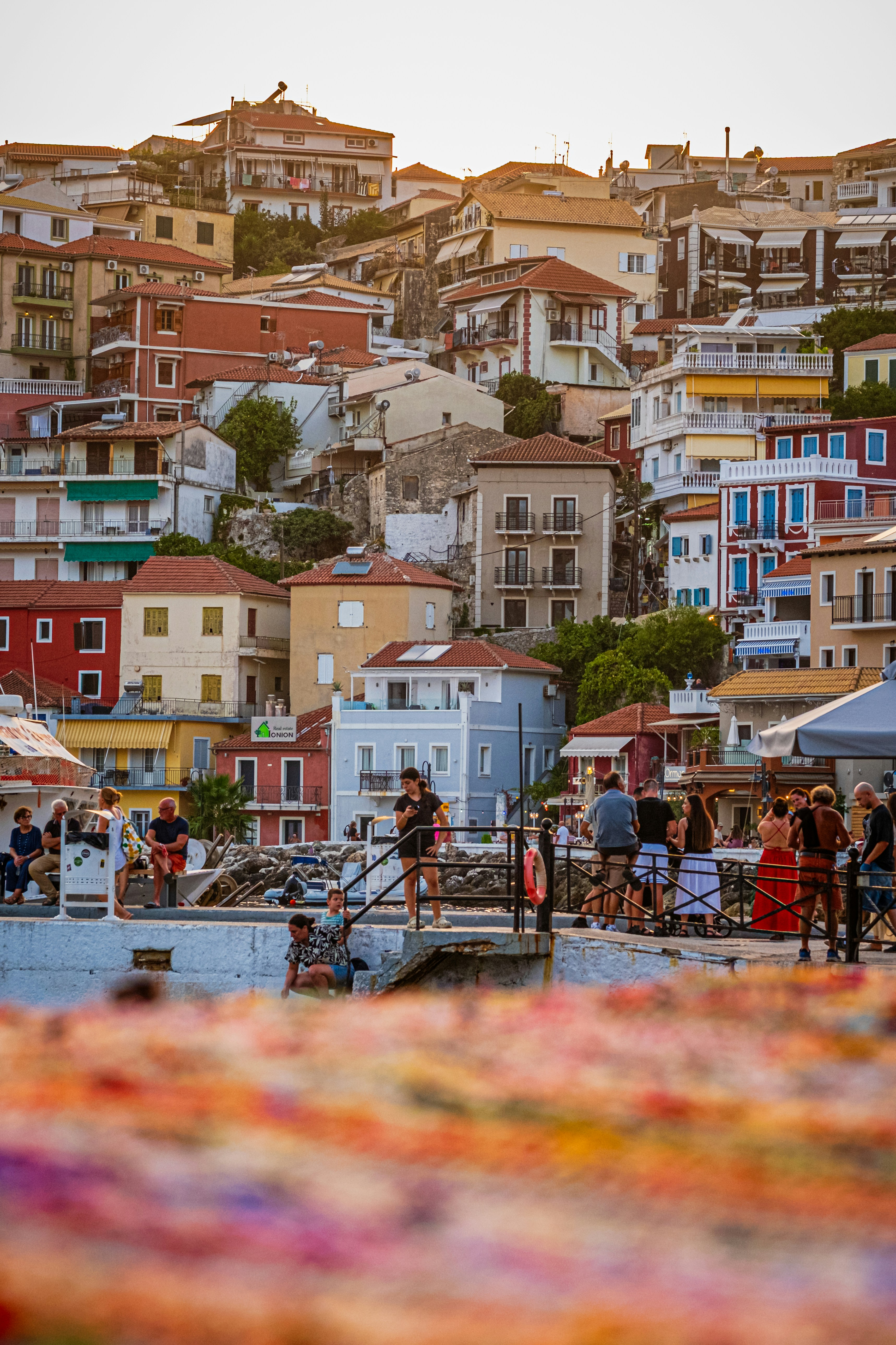 People on a harbor pier below a hillside town with colorful buildings
