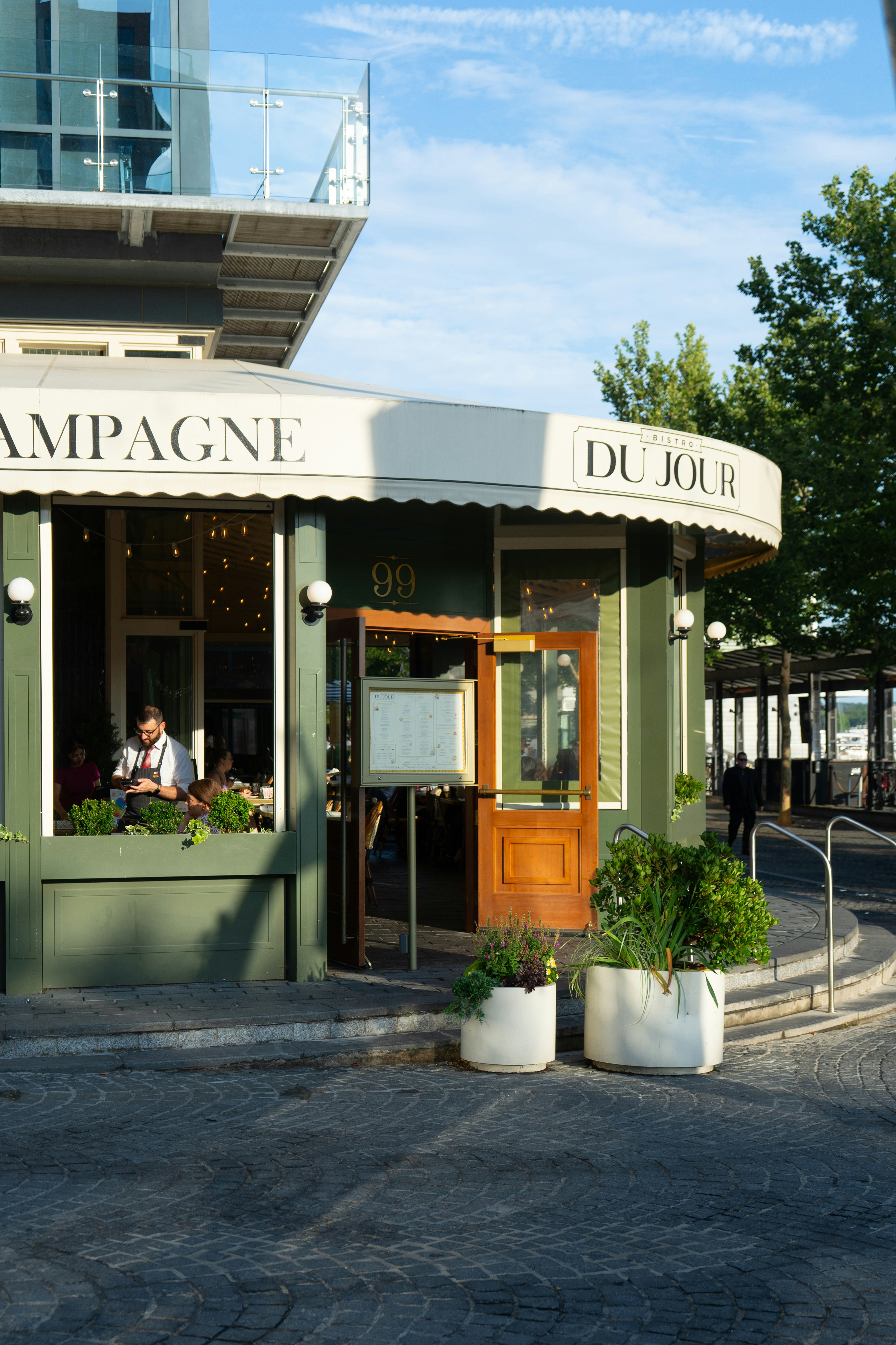 Ein grünes Bistrogeschäft mit einem weißen Vordach mit der Aufschrift "DU JOUR" und Topfpflanzen draußen