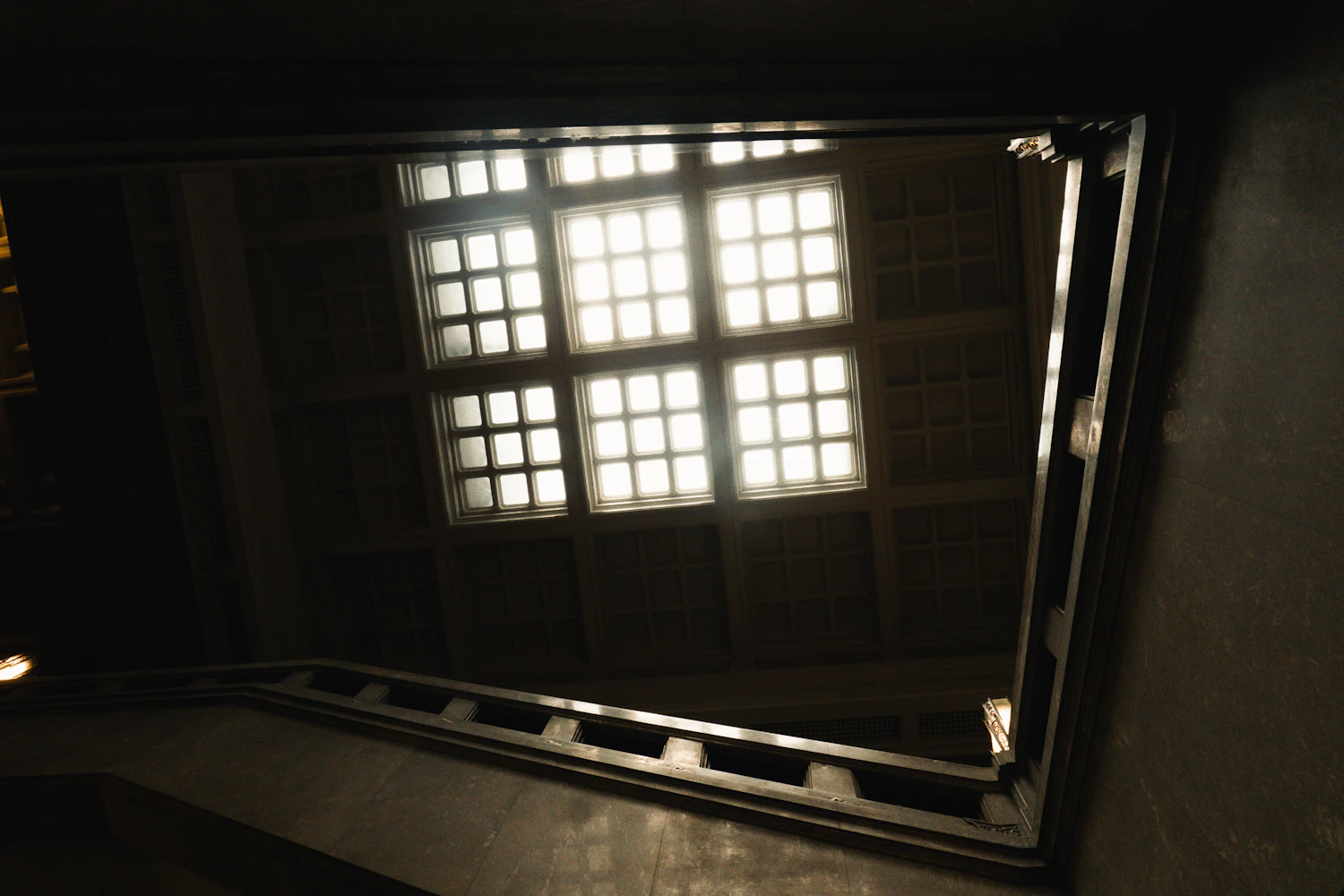 A bright gridded skylight above a dark stone stairwell, seen from the bottom of the stairs