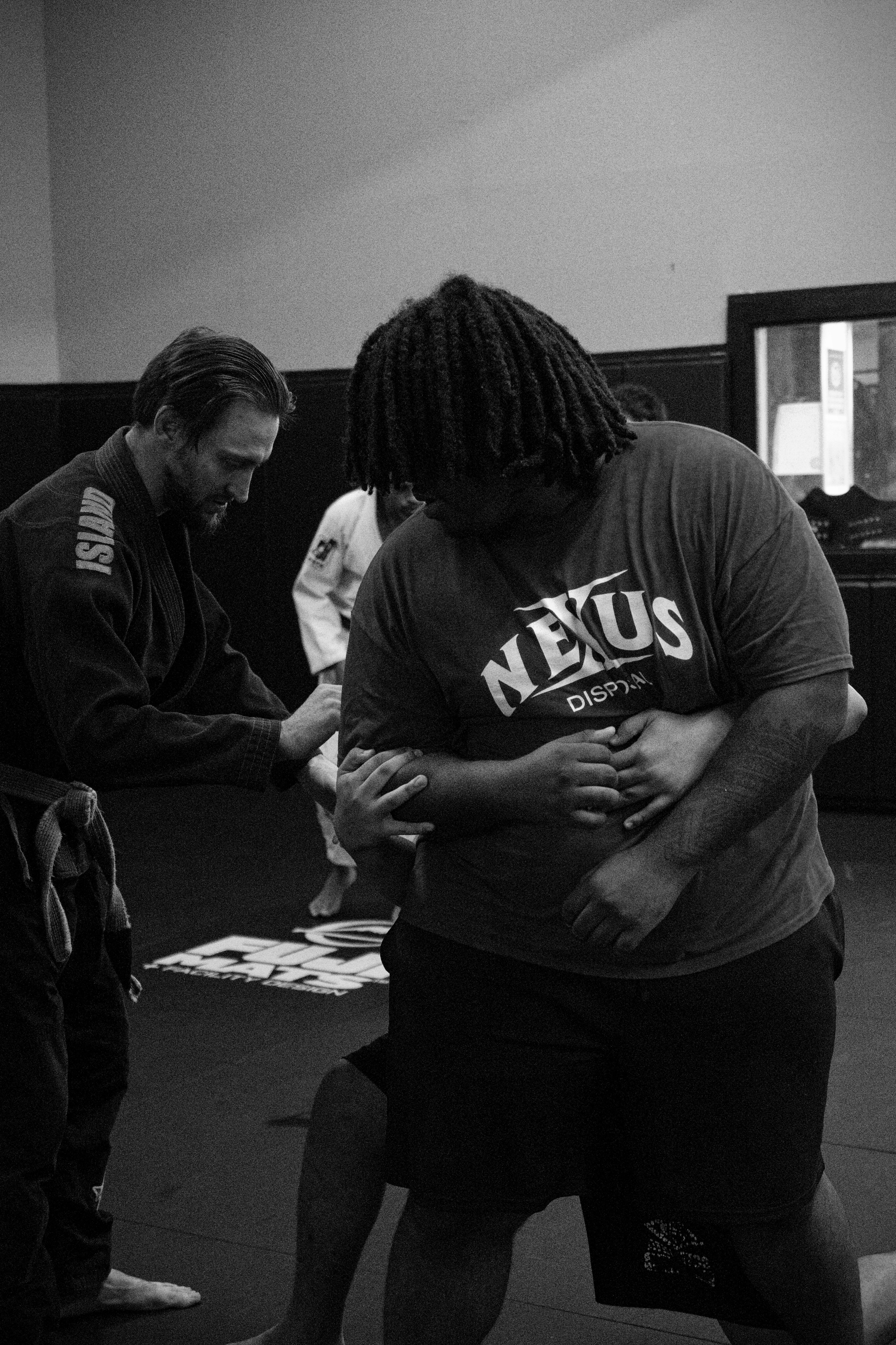 A person in a gi instructing a person in a t-shirt on a mat