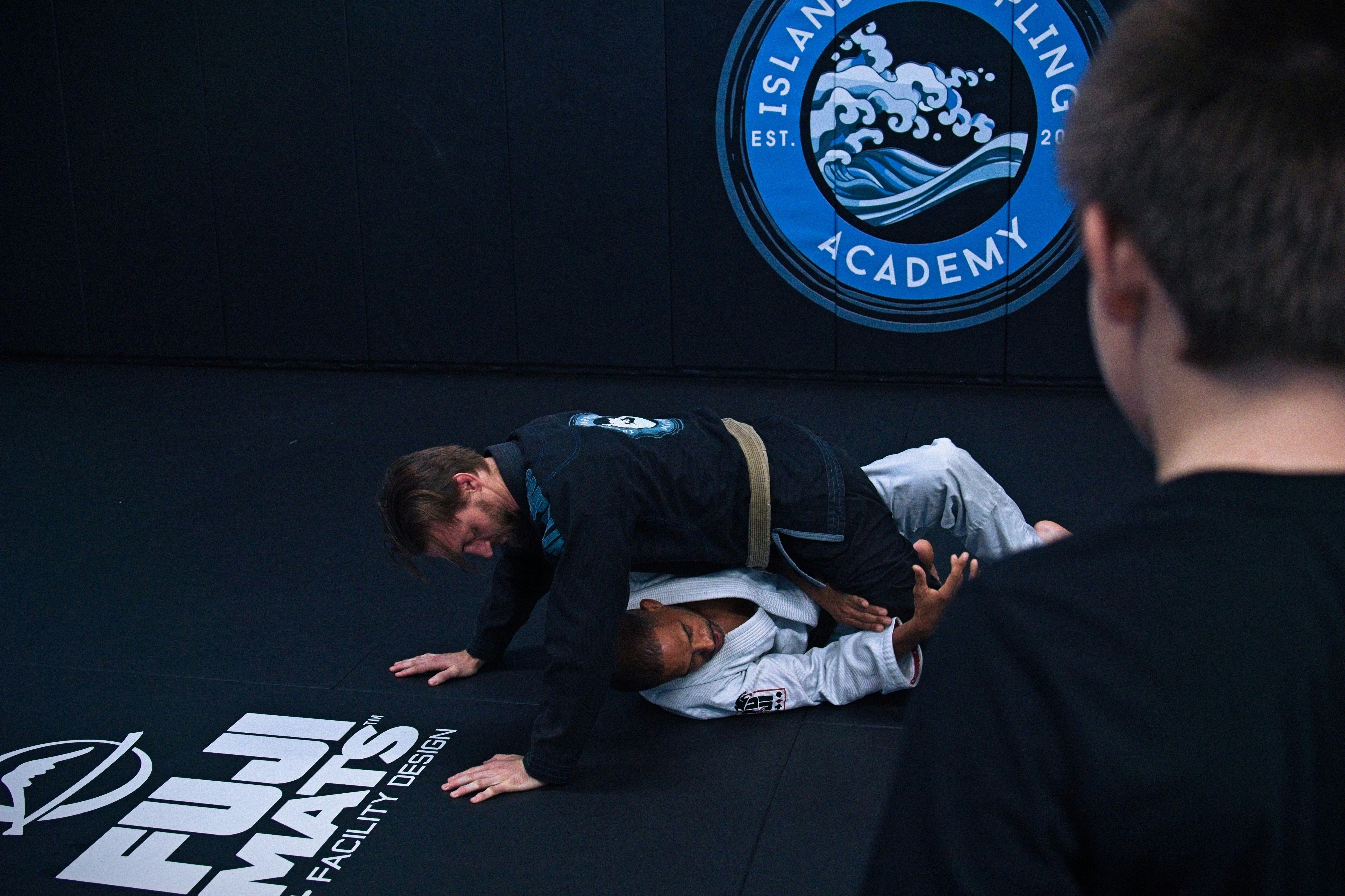 Two people in gis grappling on a black mat in a training academy