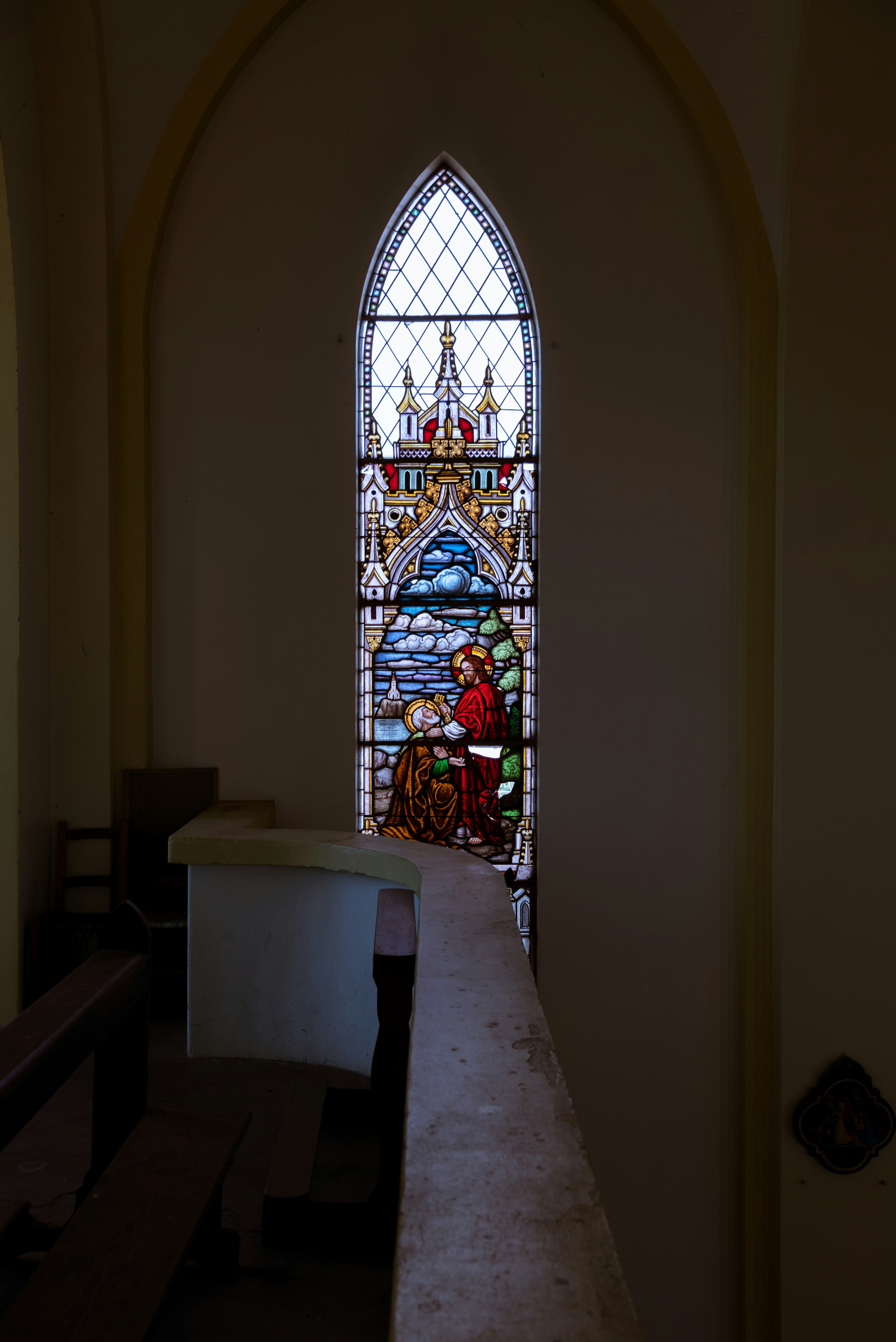 A tall arched stained glass window depicting a religious scene in a dim church