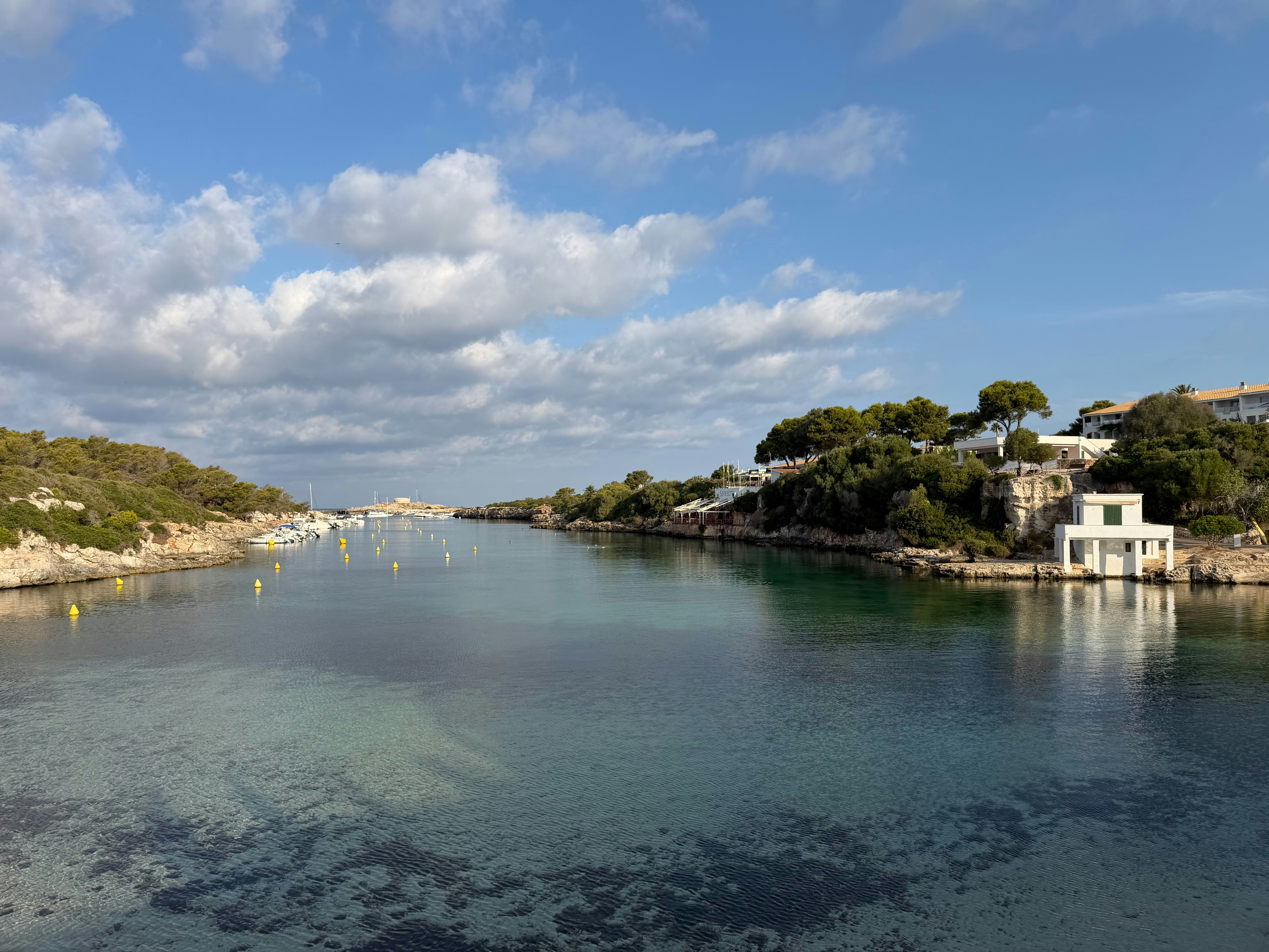 A calm coastal inlet with clear water, small boats, and rocky shores under clouds