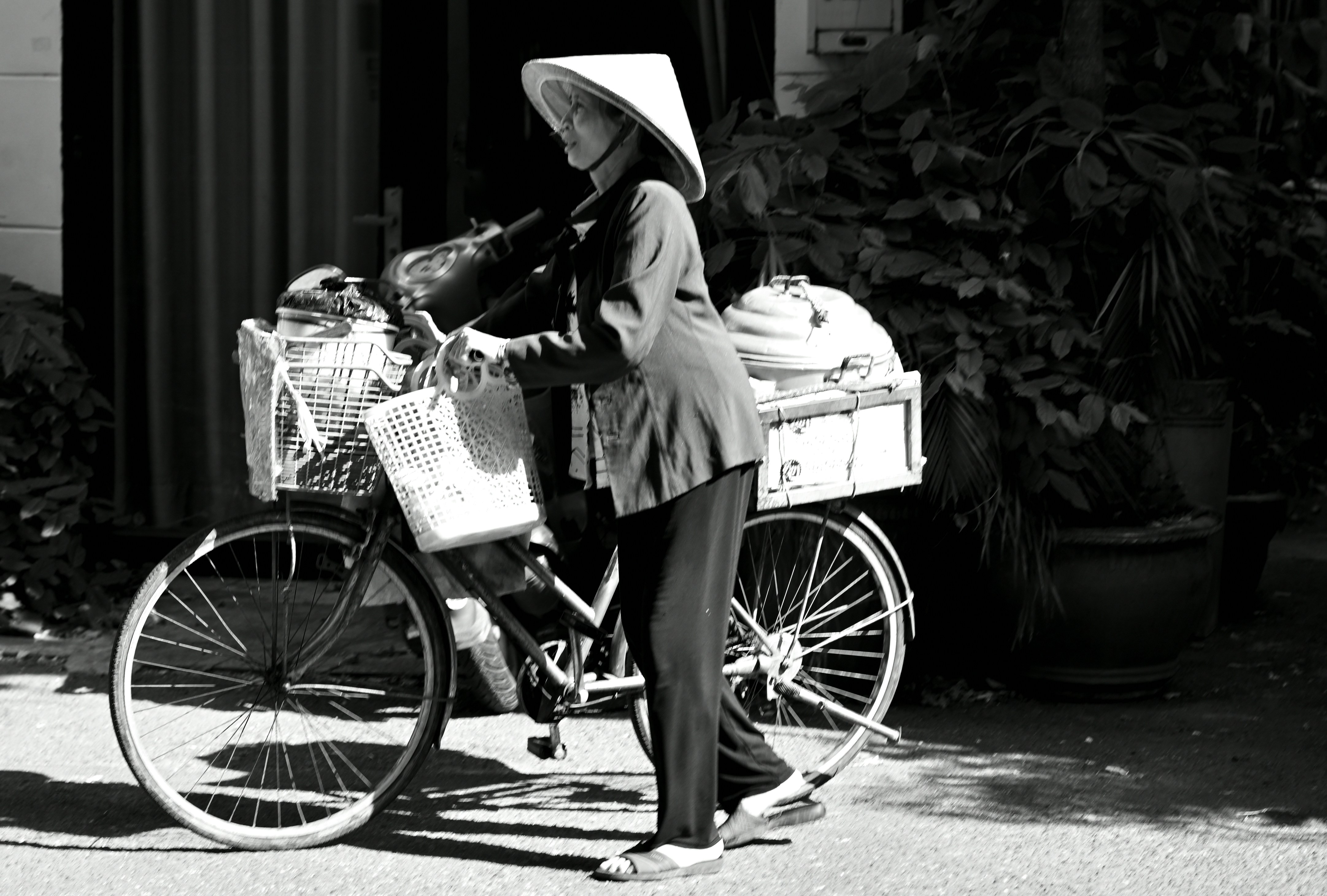 A woman in a conical hat walking a bicycle loaded with baskets and containers