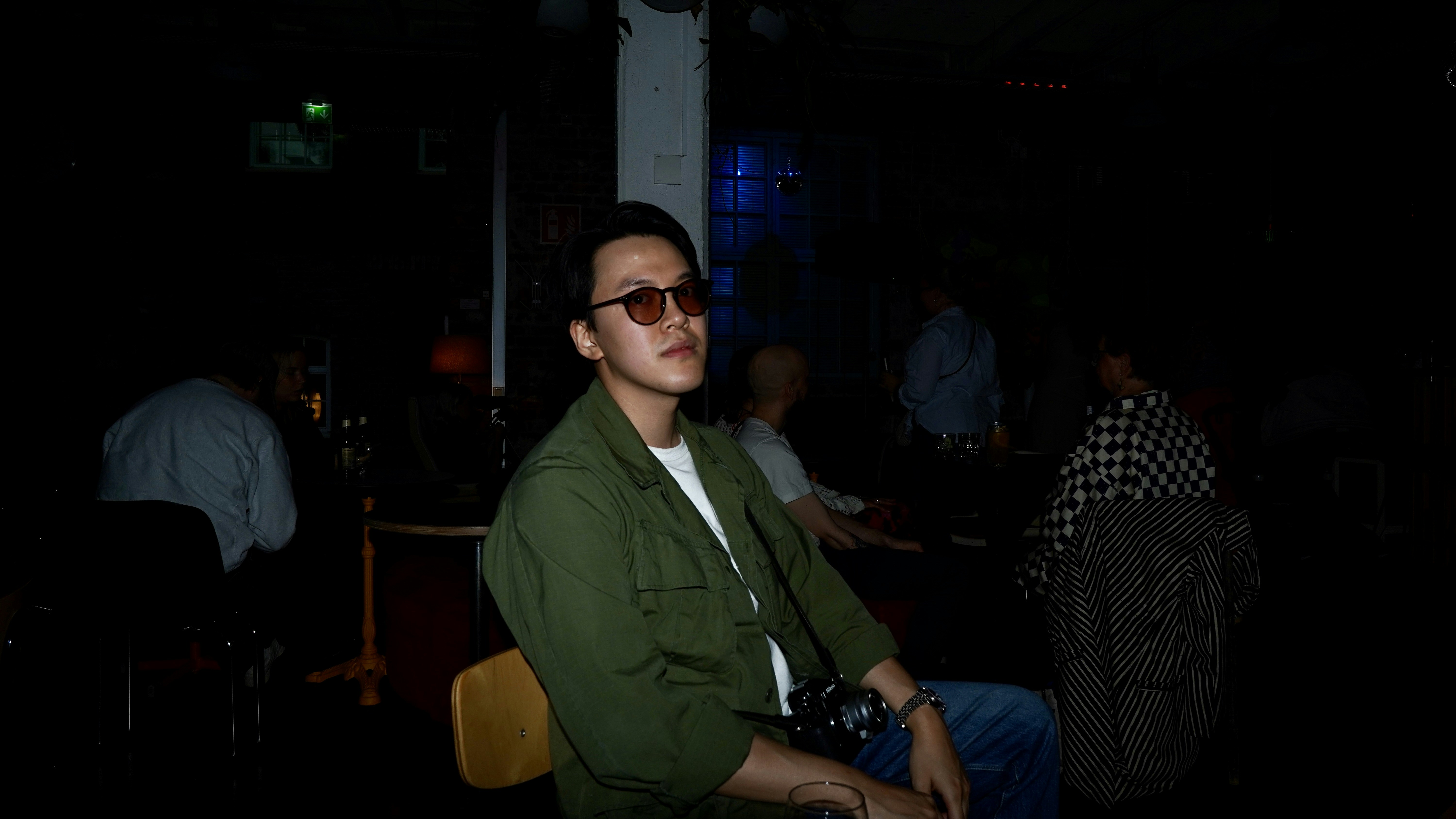 A man in a green shirt and sunglasses sitting in a dimly lit bar