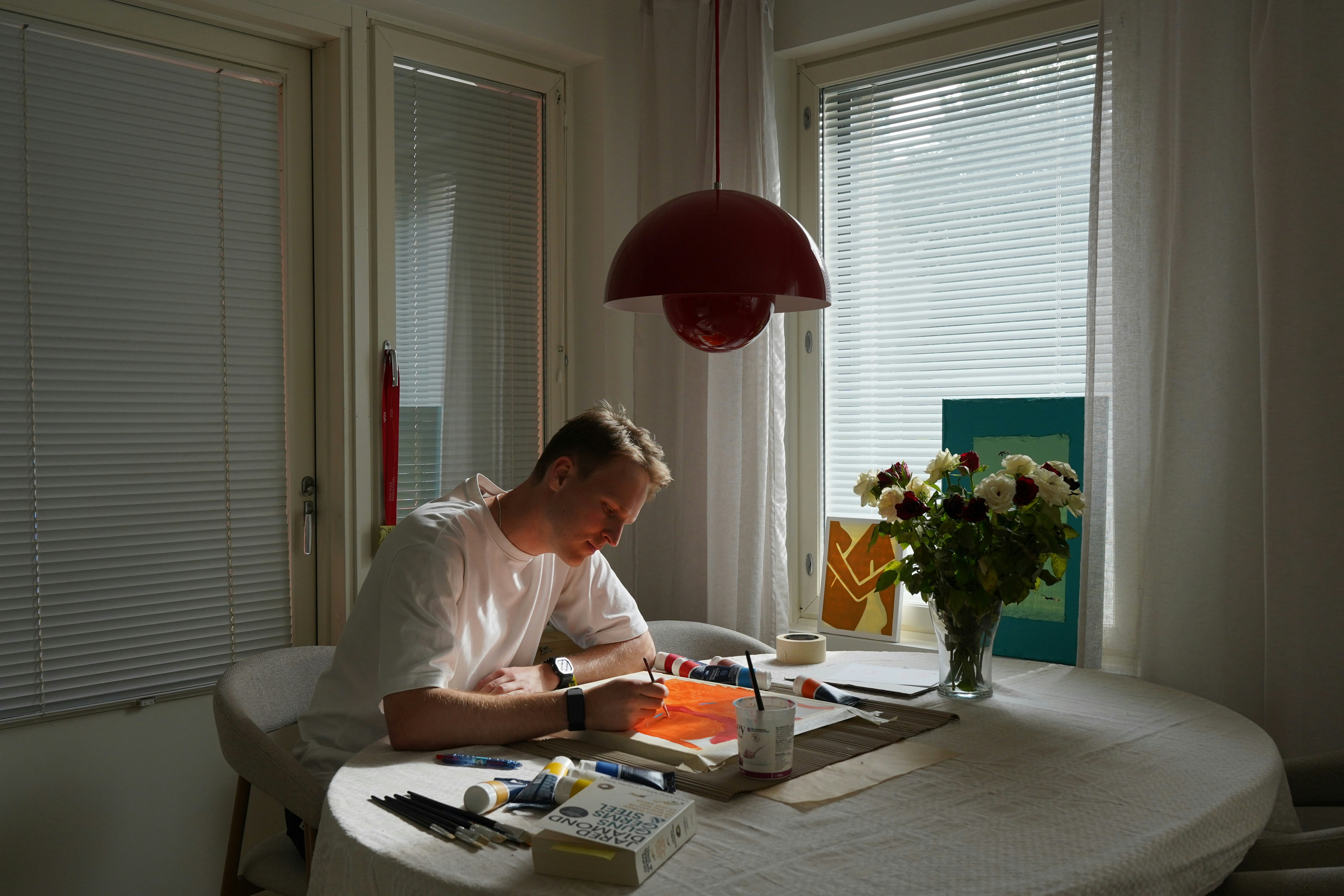 A man painting on paper at a round table with flowers in a sunlit room