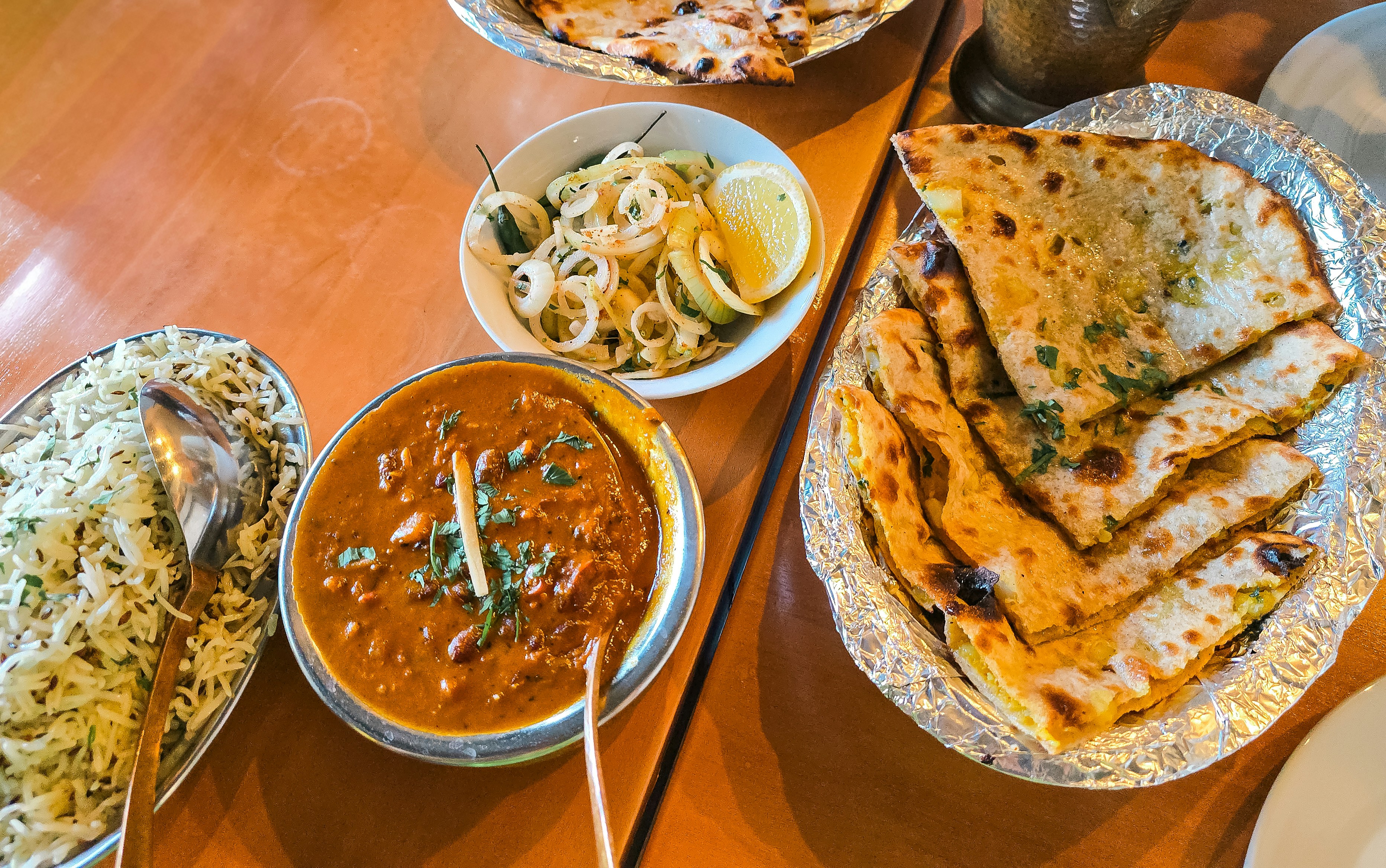 A spread from an Indian restaurant featuring a rich bowl of rajma curry, fragrant jeera rice, traditional Indian flatbreads, and a crisp, spiced onion salad.