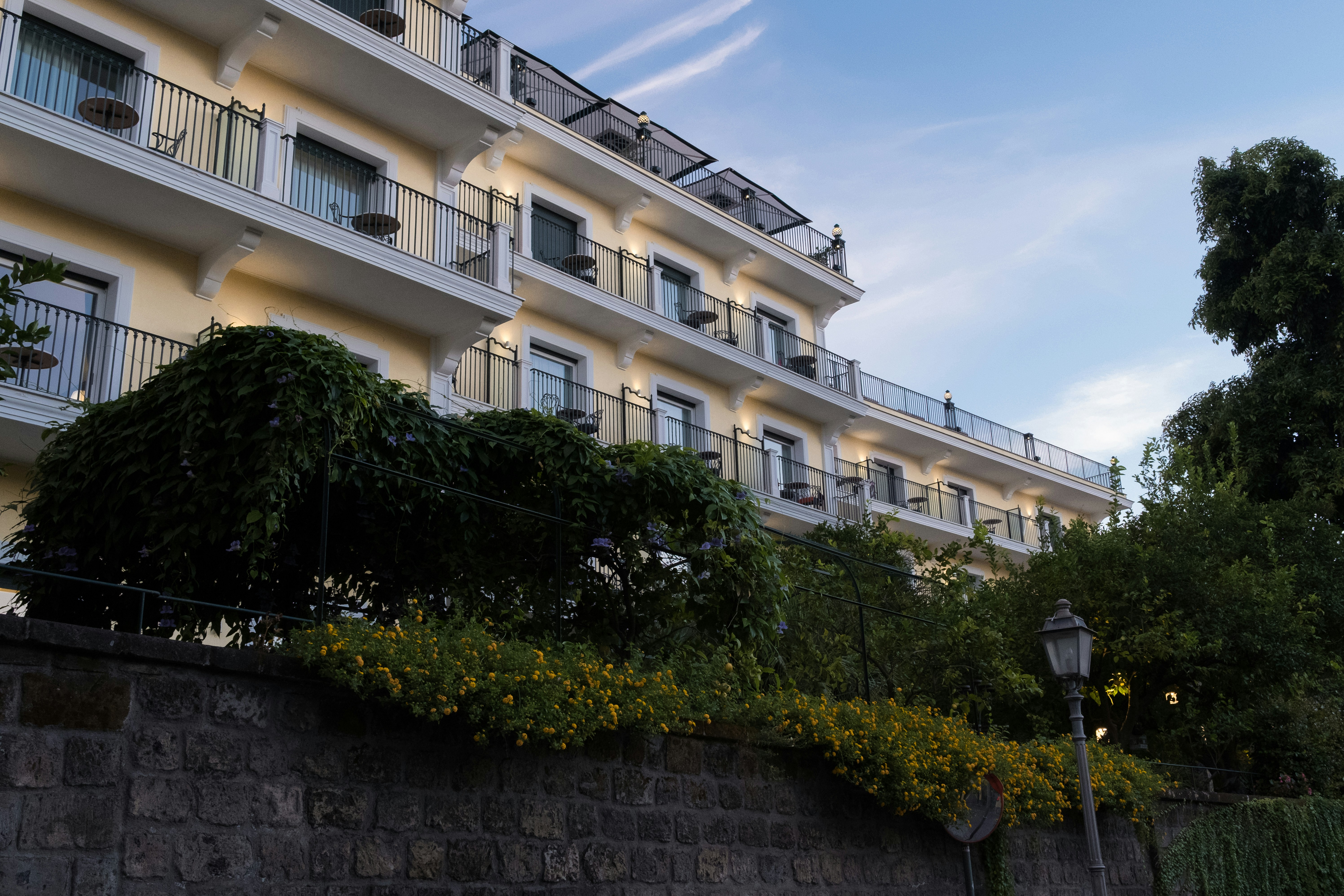 A multi-story hotel with balconies above a stone wall covered in yellow flowers