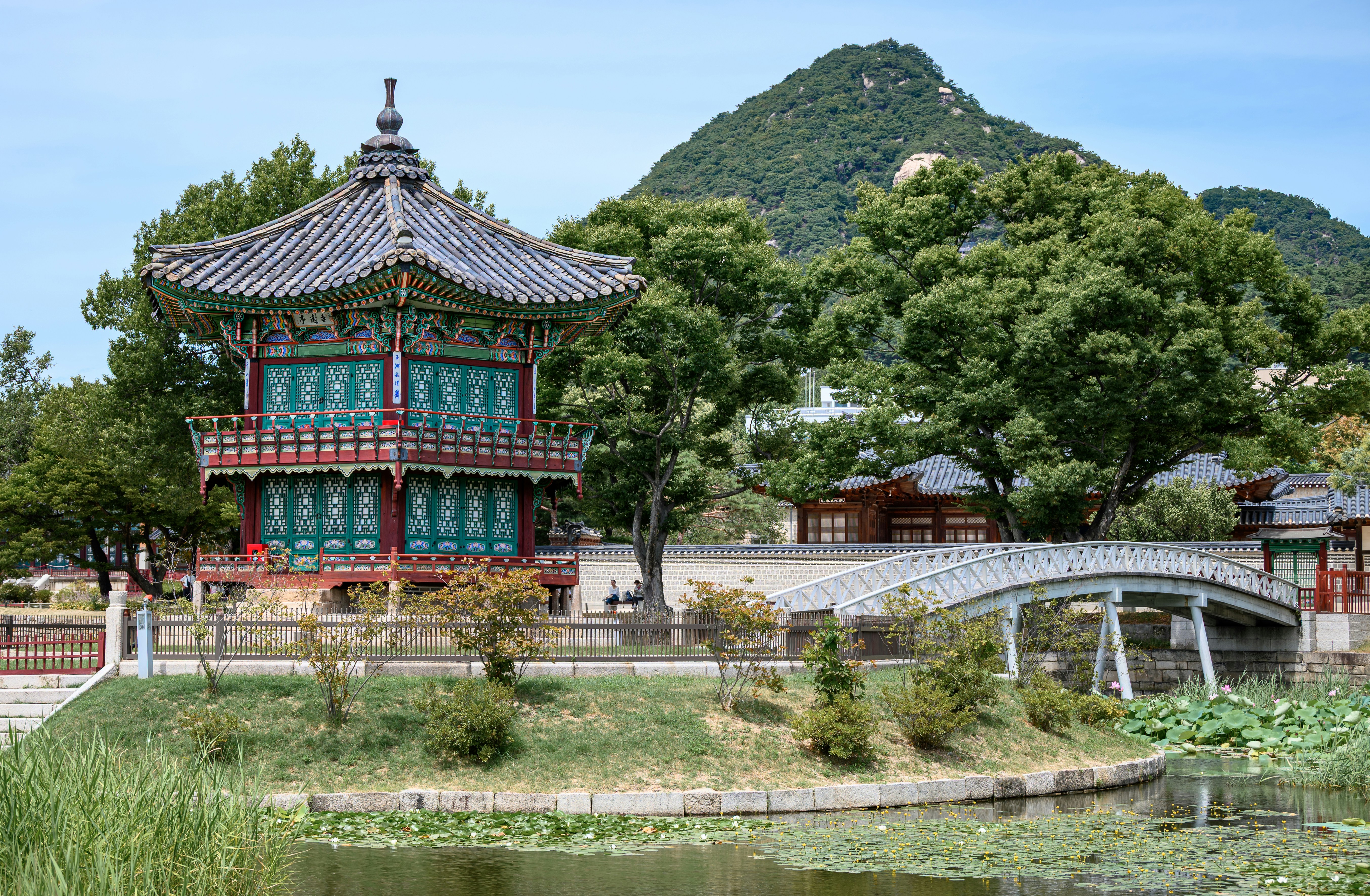 A traditional Korean pavilion beside a pond and arched bridge with a mountain background