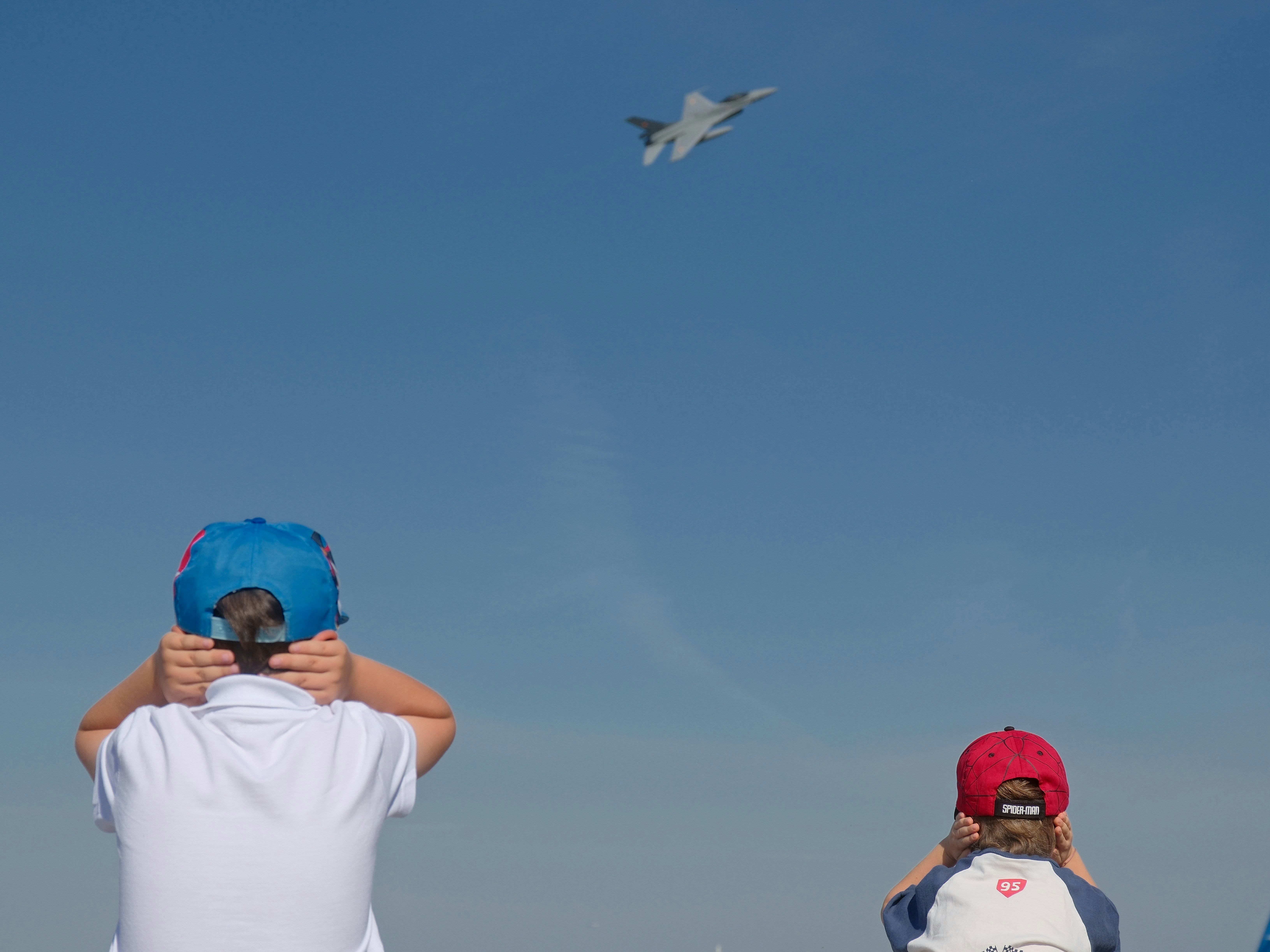 Two children in baseball caps covering their ears as a fighter jet flies overhead