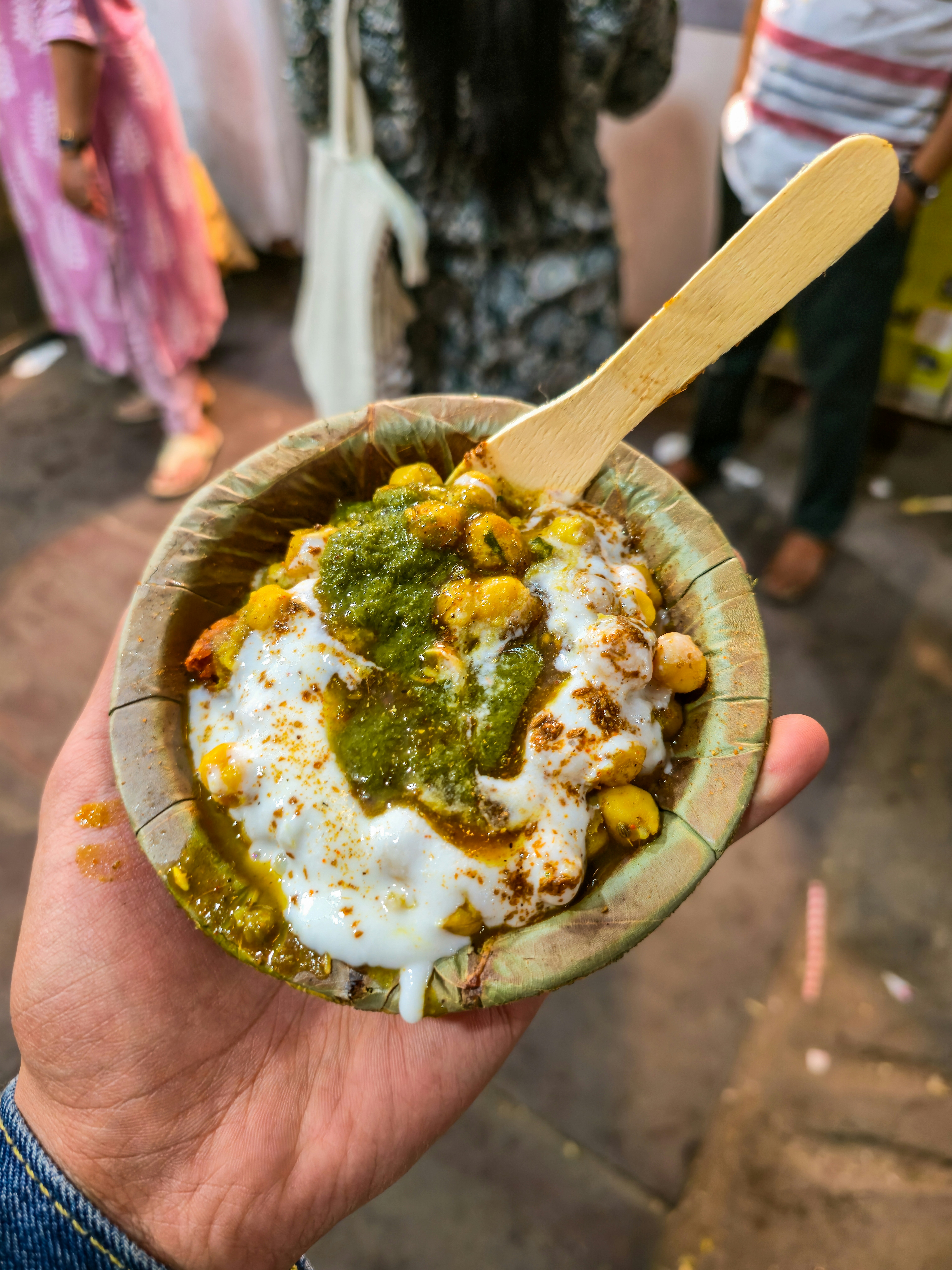 A leaf bowl of chana chaat topped with yoghurt and green chutney, held up on a busy street