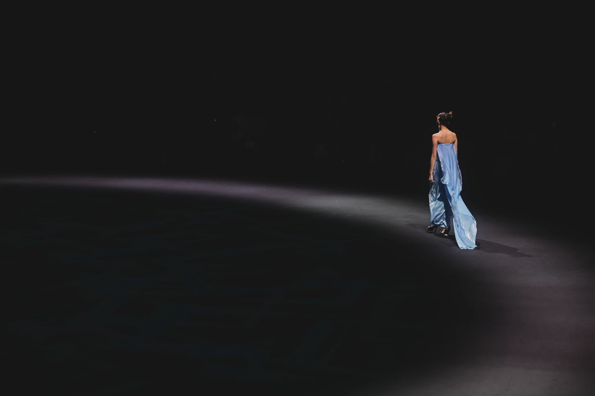 A person in a flowing light blue dress walking on a dark runway
