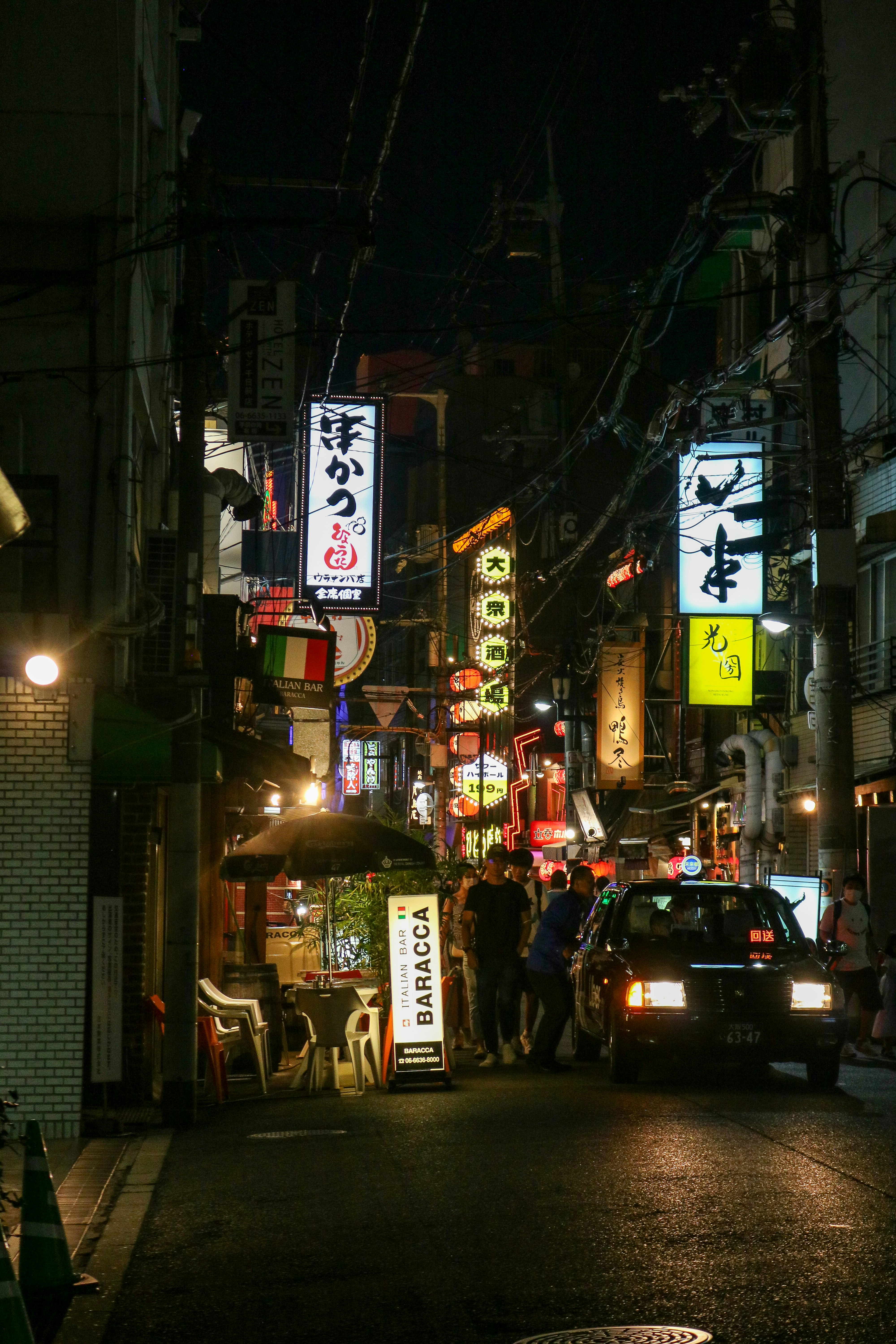 A black taxi driving down a narrow street lined with illuminated Japanese signs
