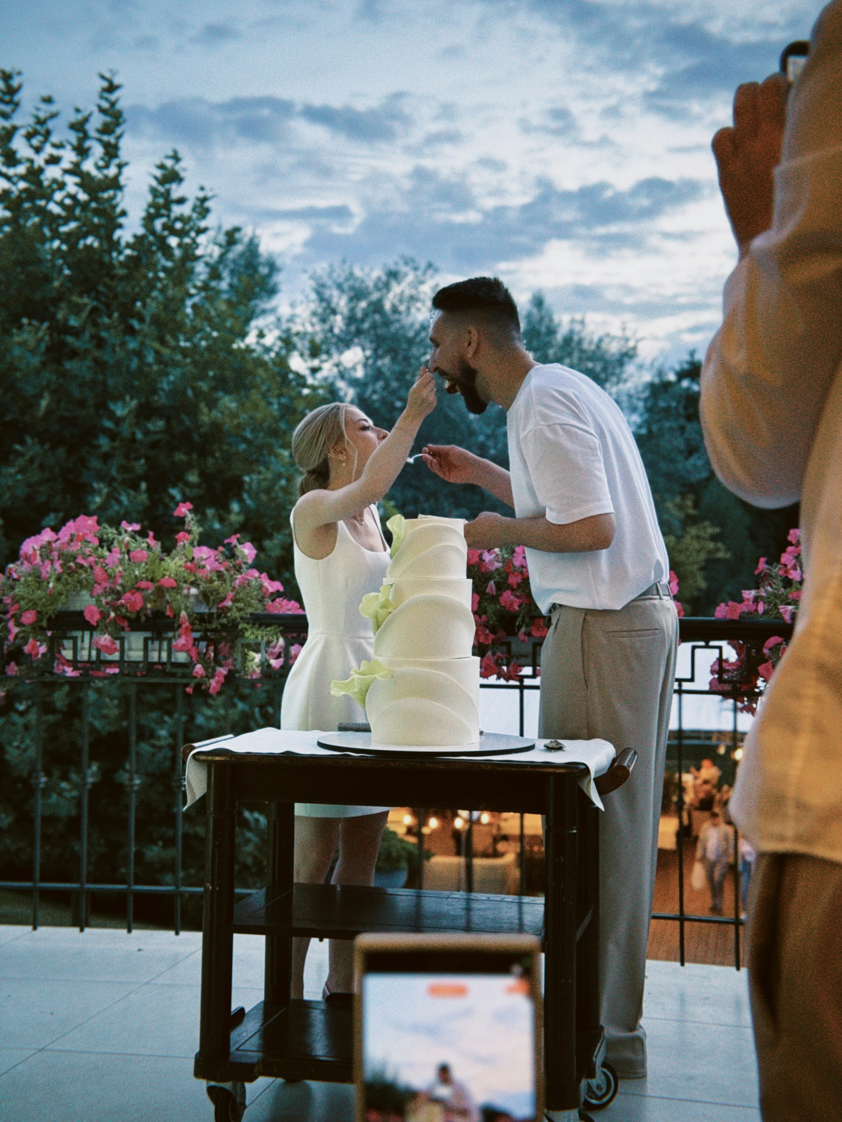 A woman in a white dress feeding cake to a man outdoors