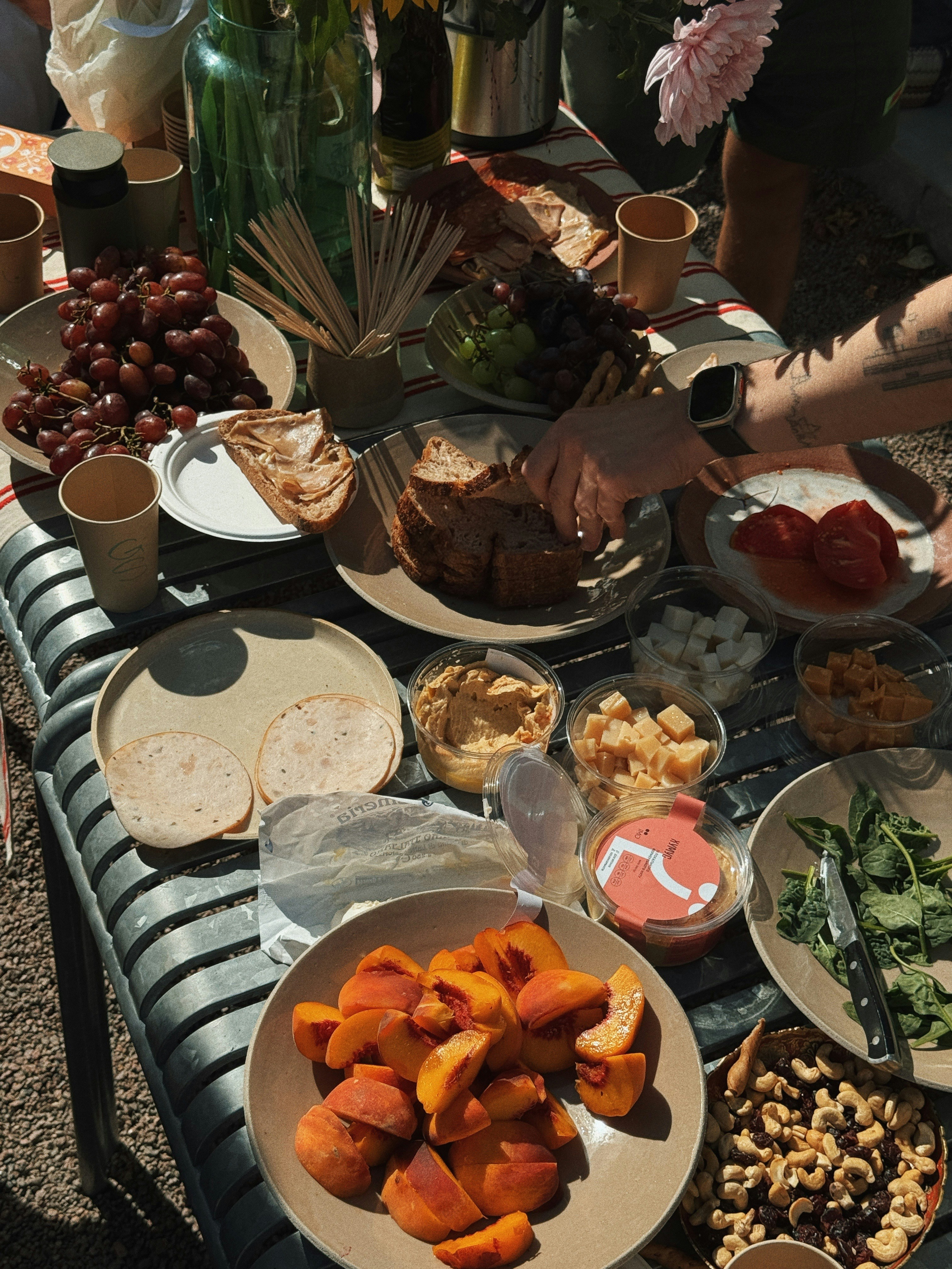 A person reaching for bread on a table spread with fruit, cheese, and snacks
