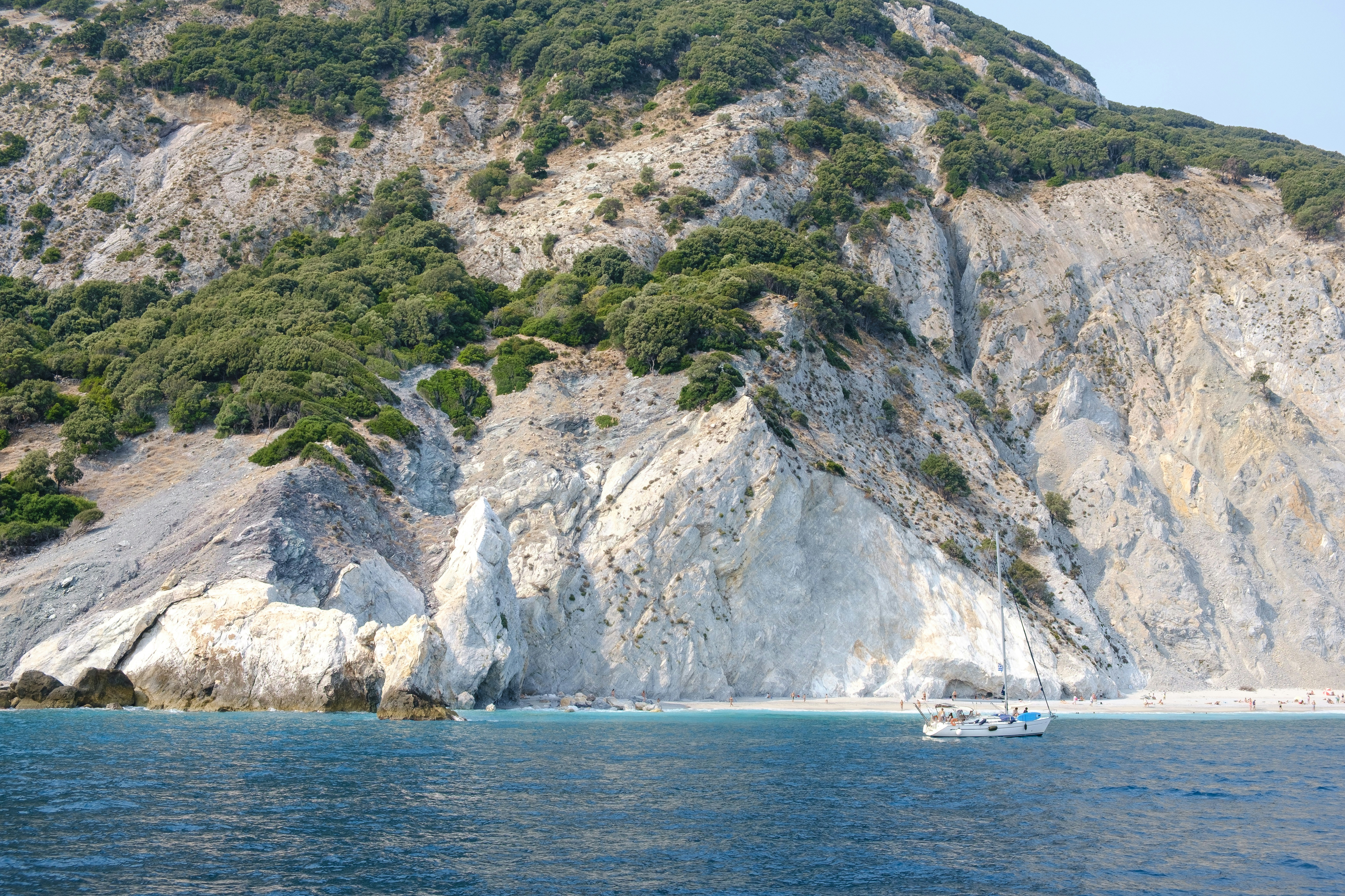 Ein Segelboot ankert nahe einer steilen, felsigen Klippe mit grünen Bäumen über dem Strand