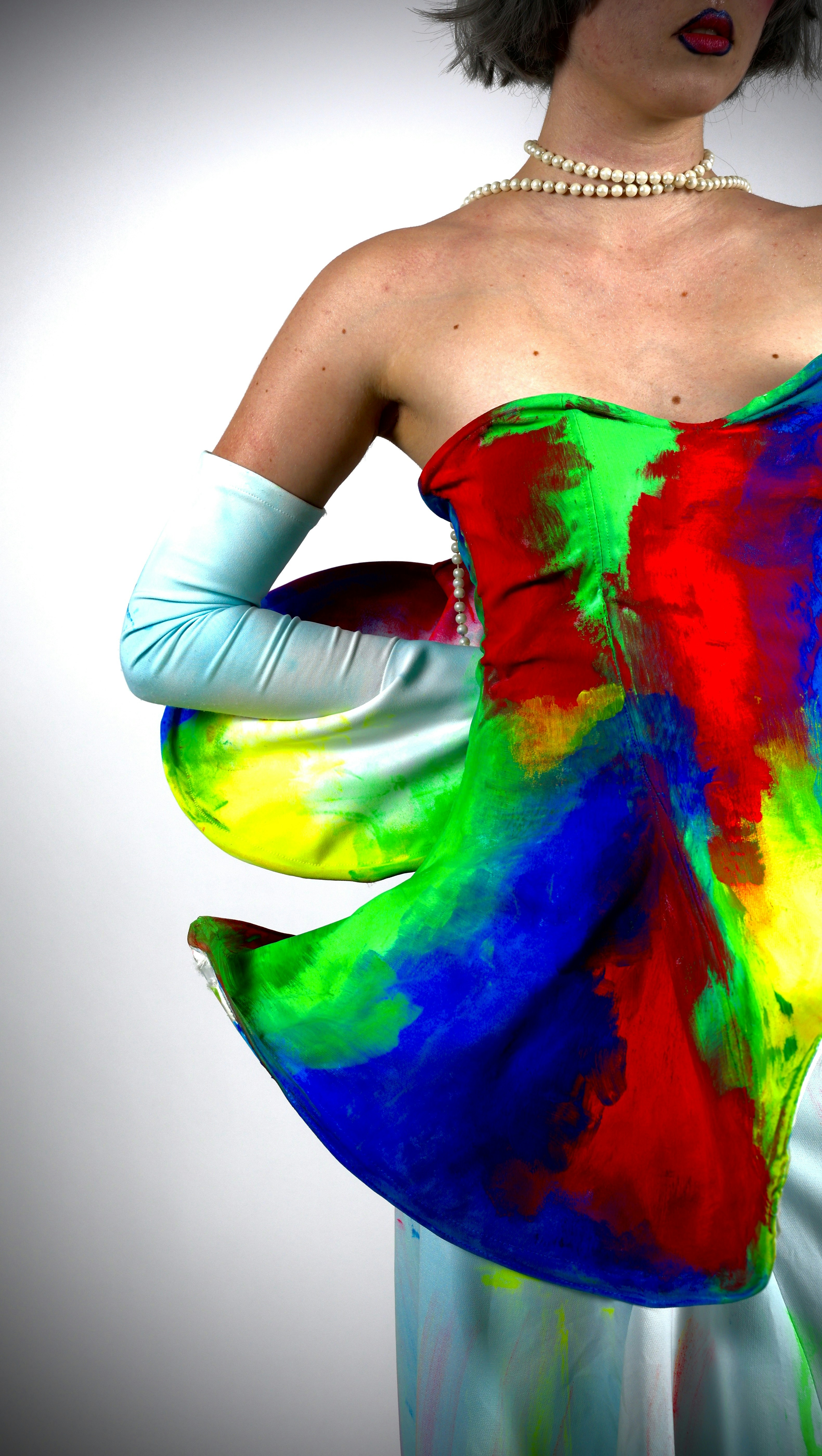 A person wearing a vibrant paint-splattered dress and light blue gloves with a pearl necklace
