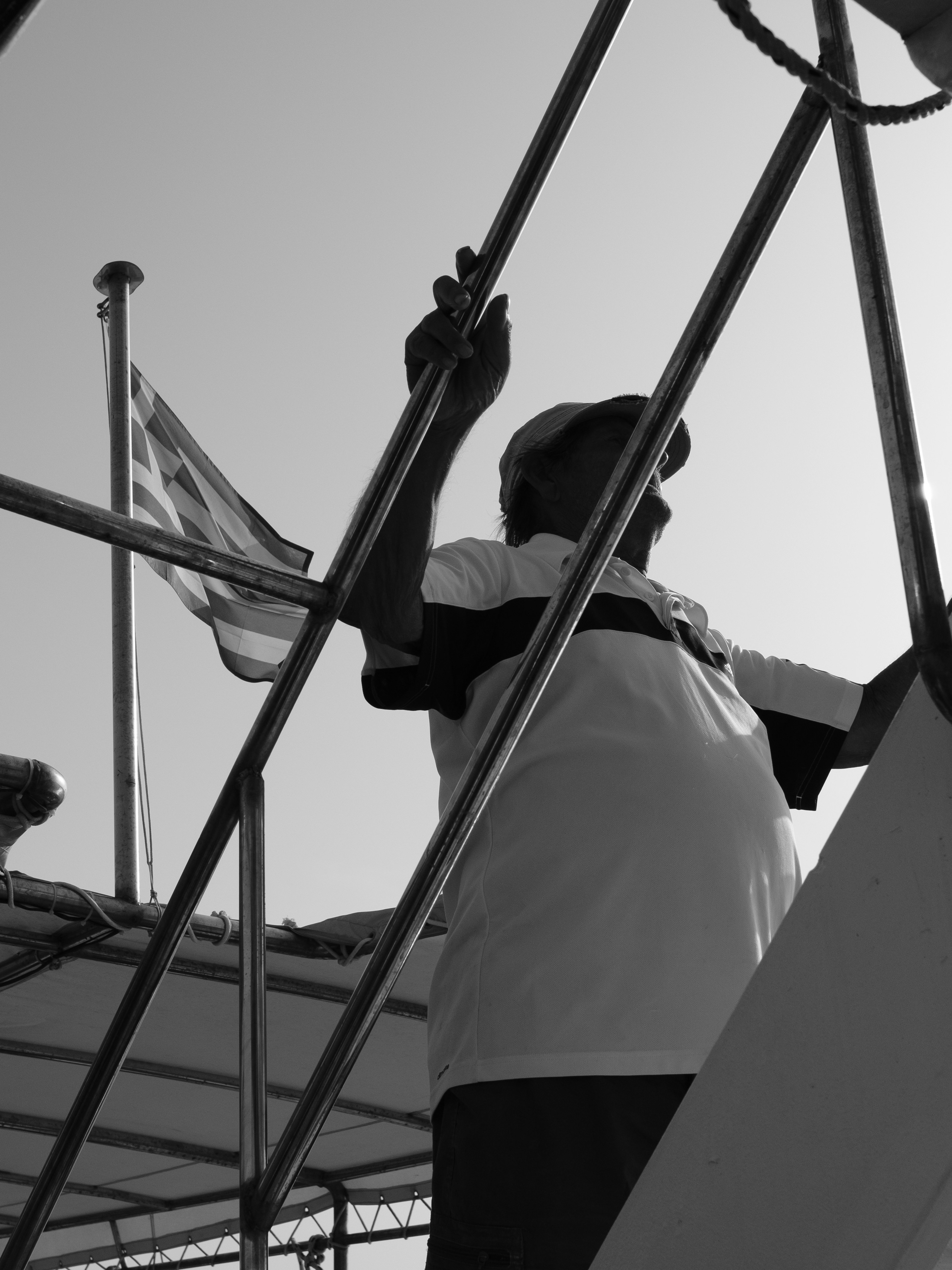 Ein Mann mit Mütze, der Metallgeländer auf einem Boot unter griechischer Flagge hält