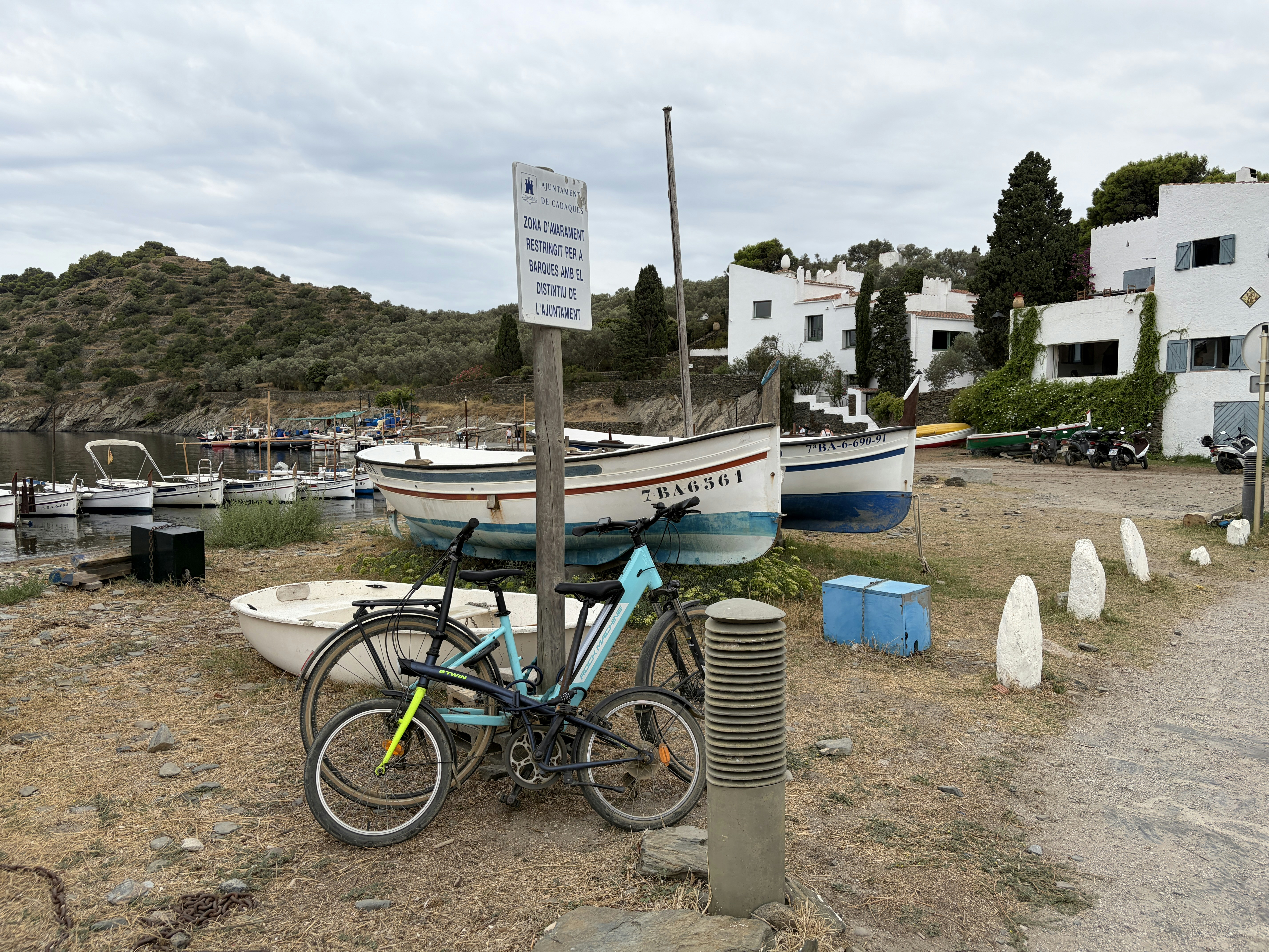 Zwei Fahrräder stehen bei einem Schild und Boote an einem felsigen Ufer in Cadaqués
