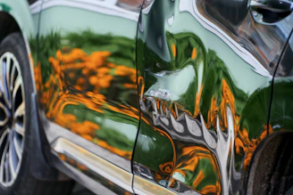 A dented dark green car door reflecting orange flowers and green foliage