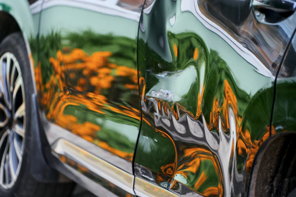 A dented dark green car door reflecting orange flowers and green foliage
