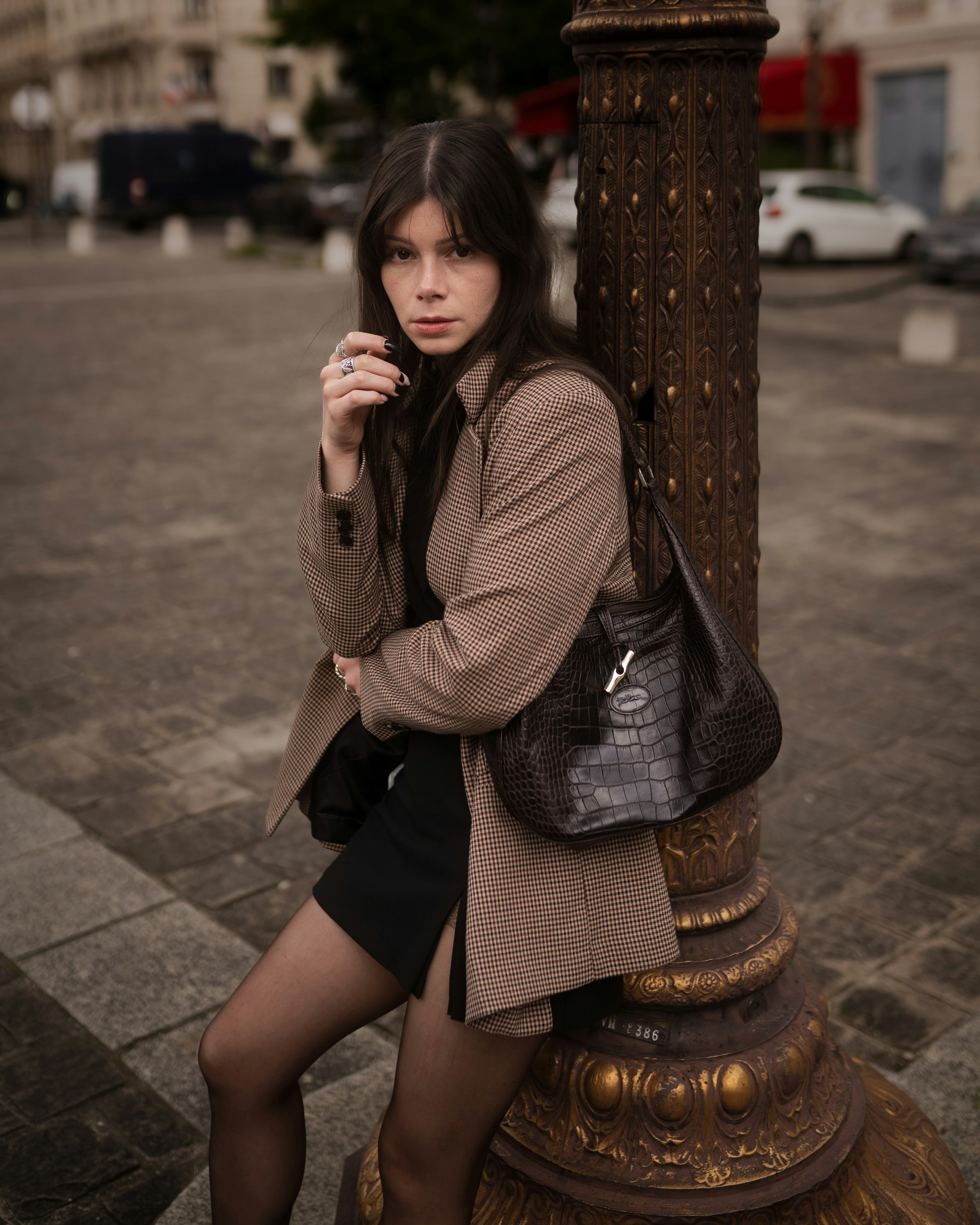 A woman in a plaid blazer and black skirt leaning against an ornate lamppost
