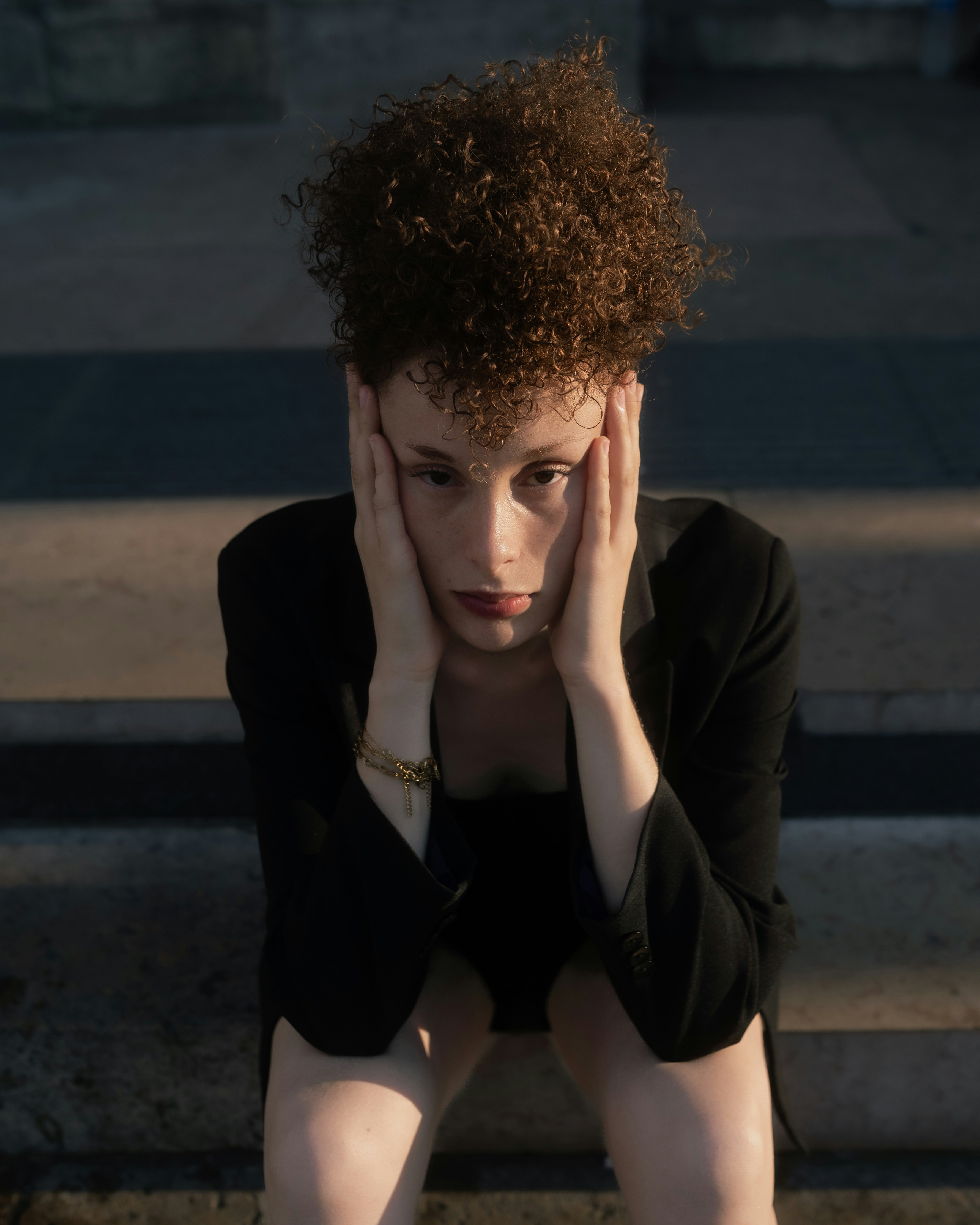 A person with curly hair holding their face in their hands while sitting outdoors