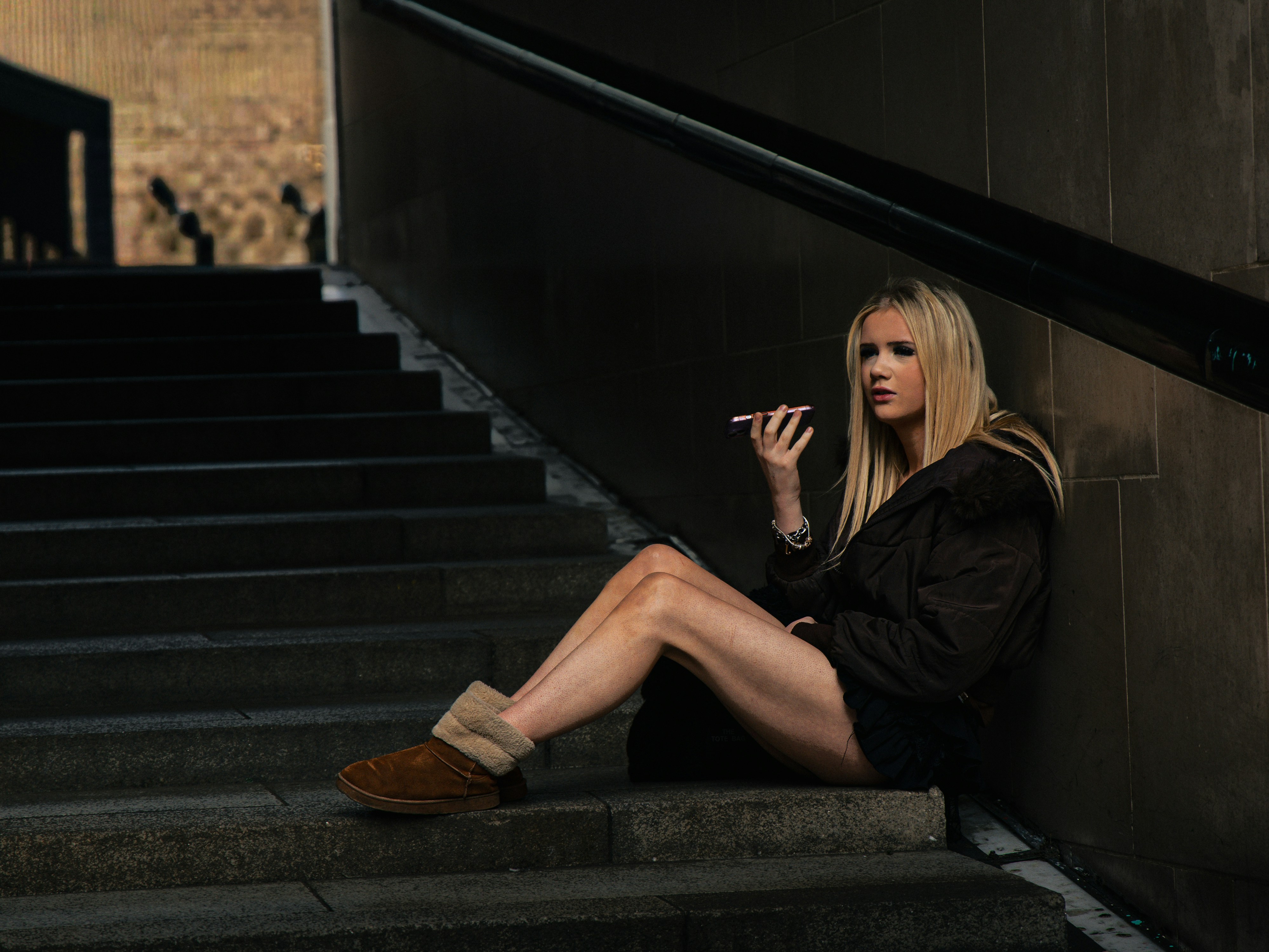 A blonde woman in a brown jacket talking on a phone on stone stairs