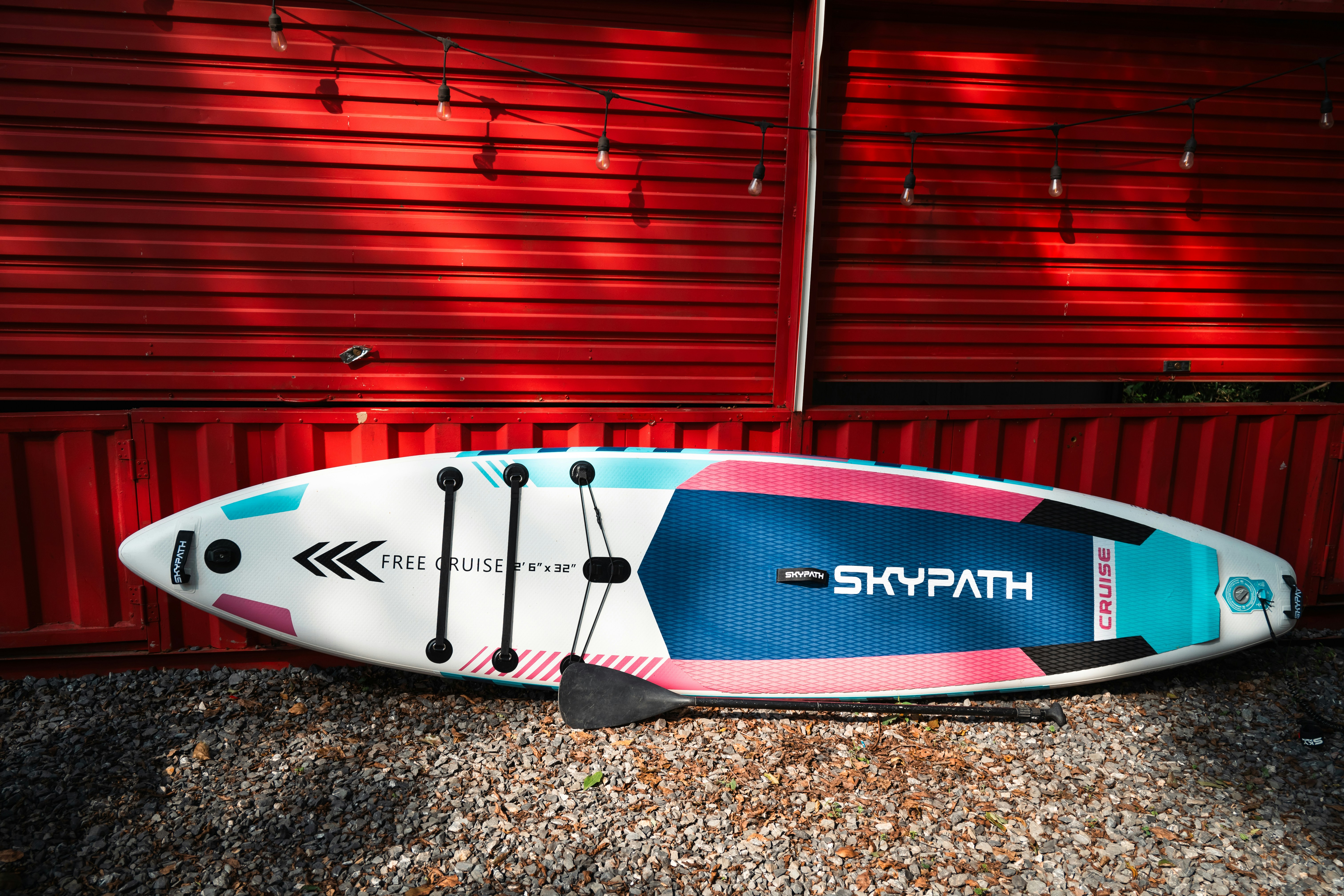 A white paddle board with blue and pink accents leaning against a red wall