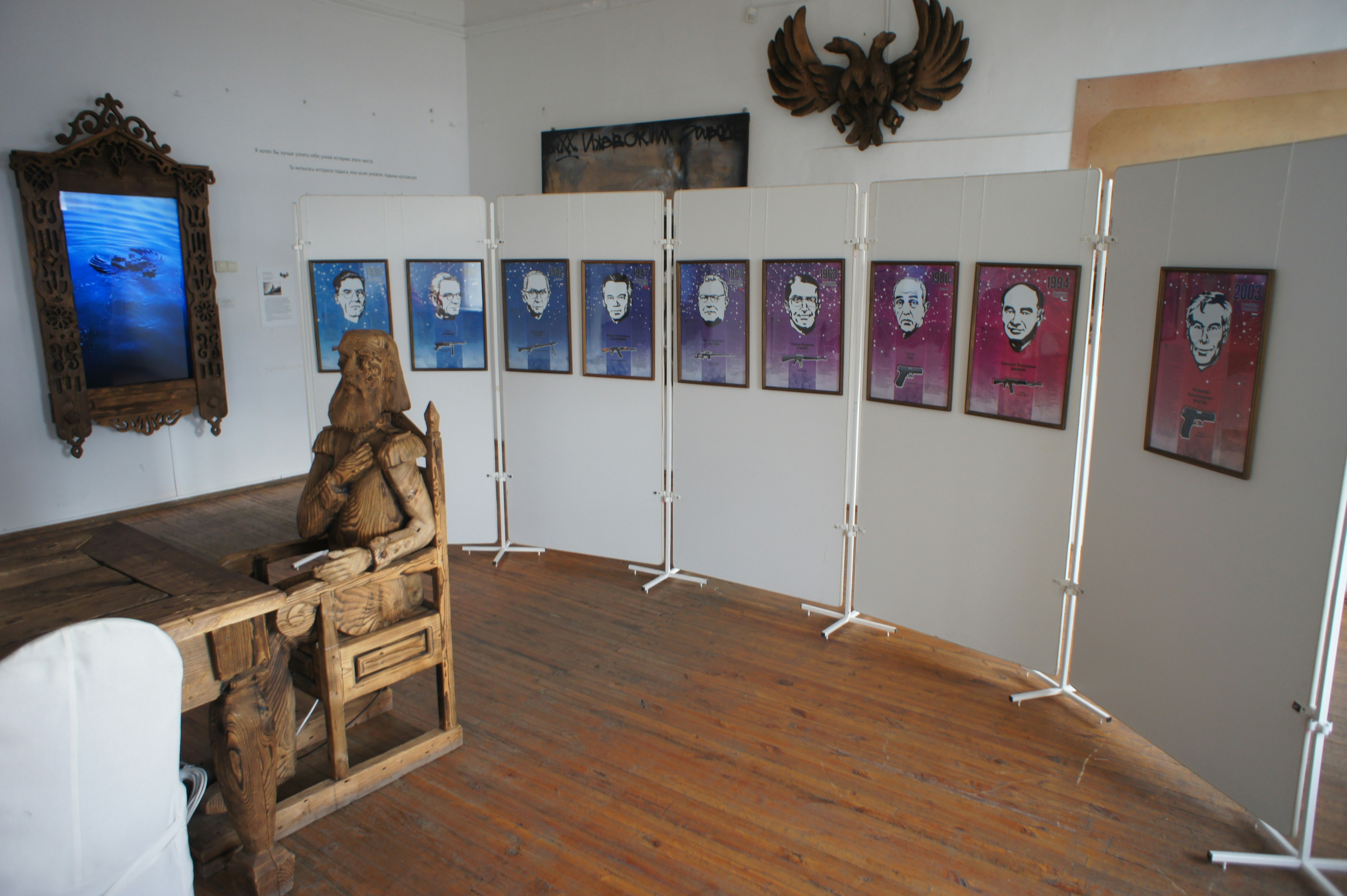 A wooden sculpture of a seated figure facing framed portraits on white display panels