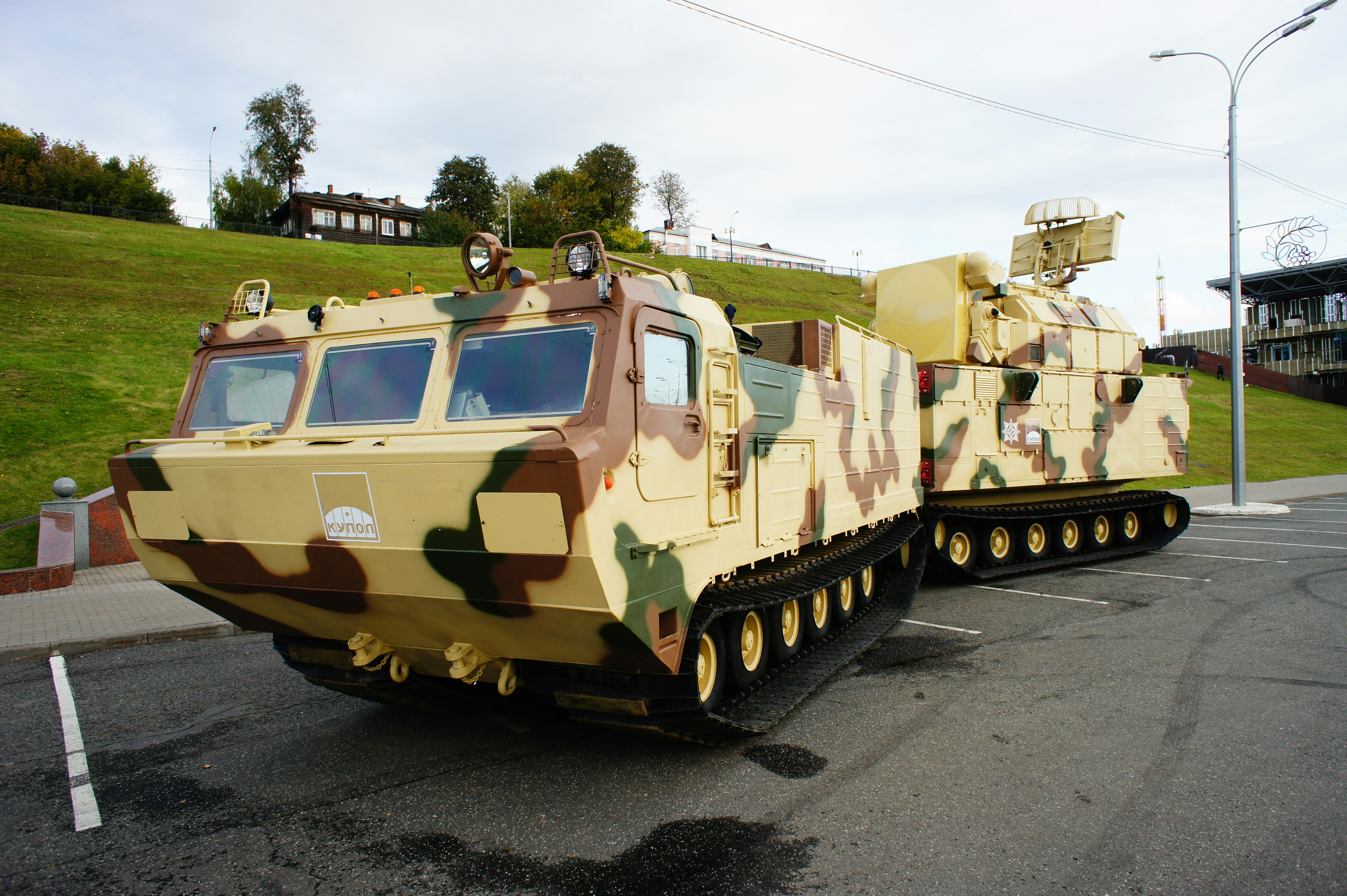A camouflaged tracked military vehicle with a radar unit parked on asphalt