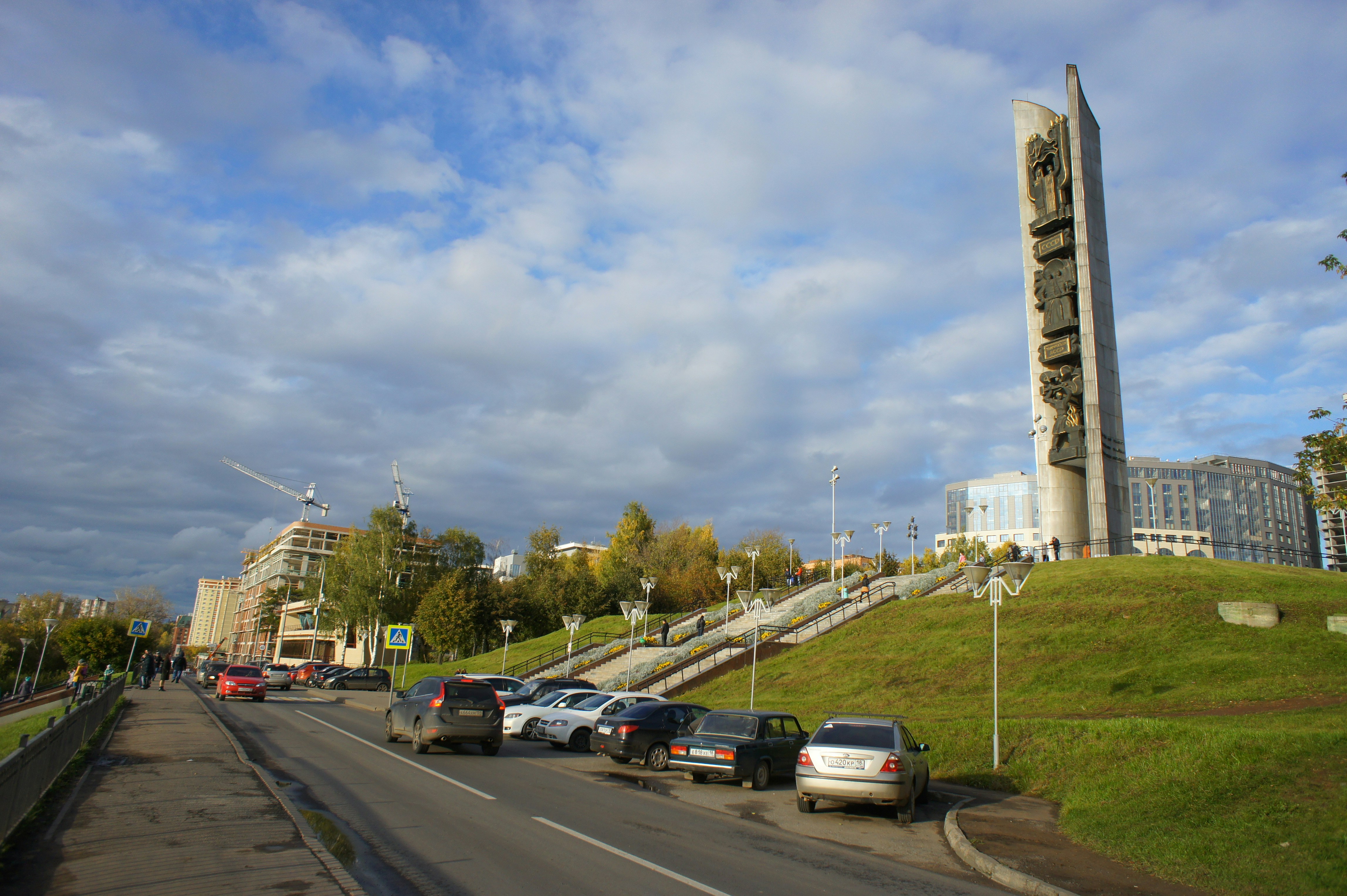 A tall concrete monument on a grassy hill above a street with parked cars