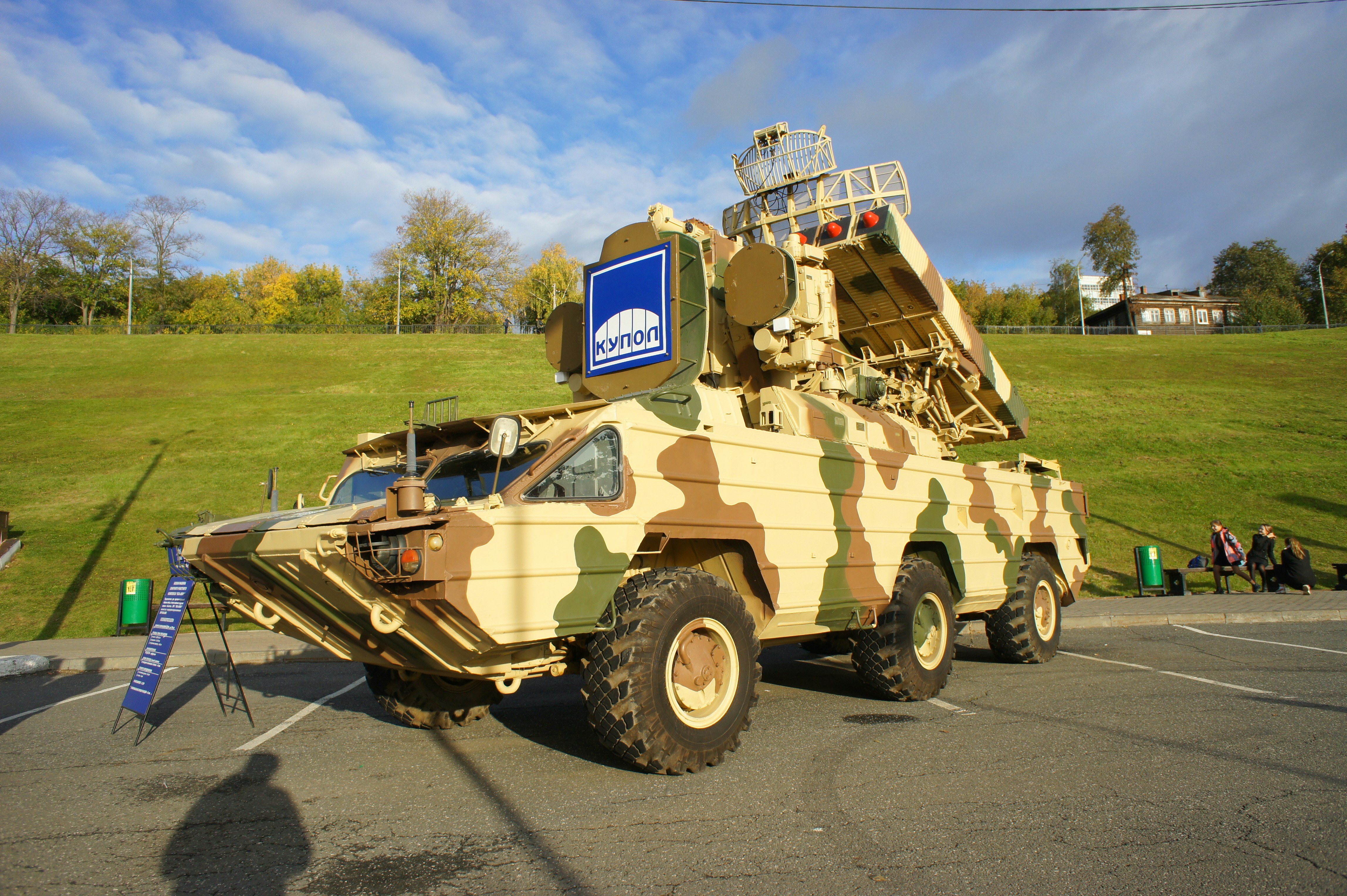 A camouflaged military air defense vehicle parked on an asphalt lot near a grassy hill
