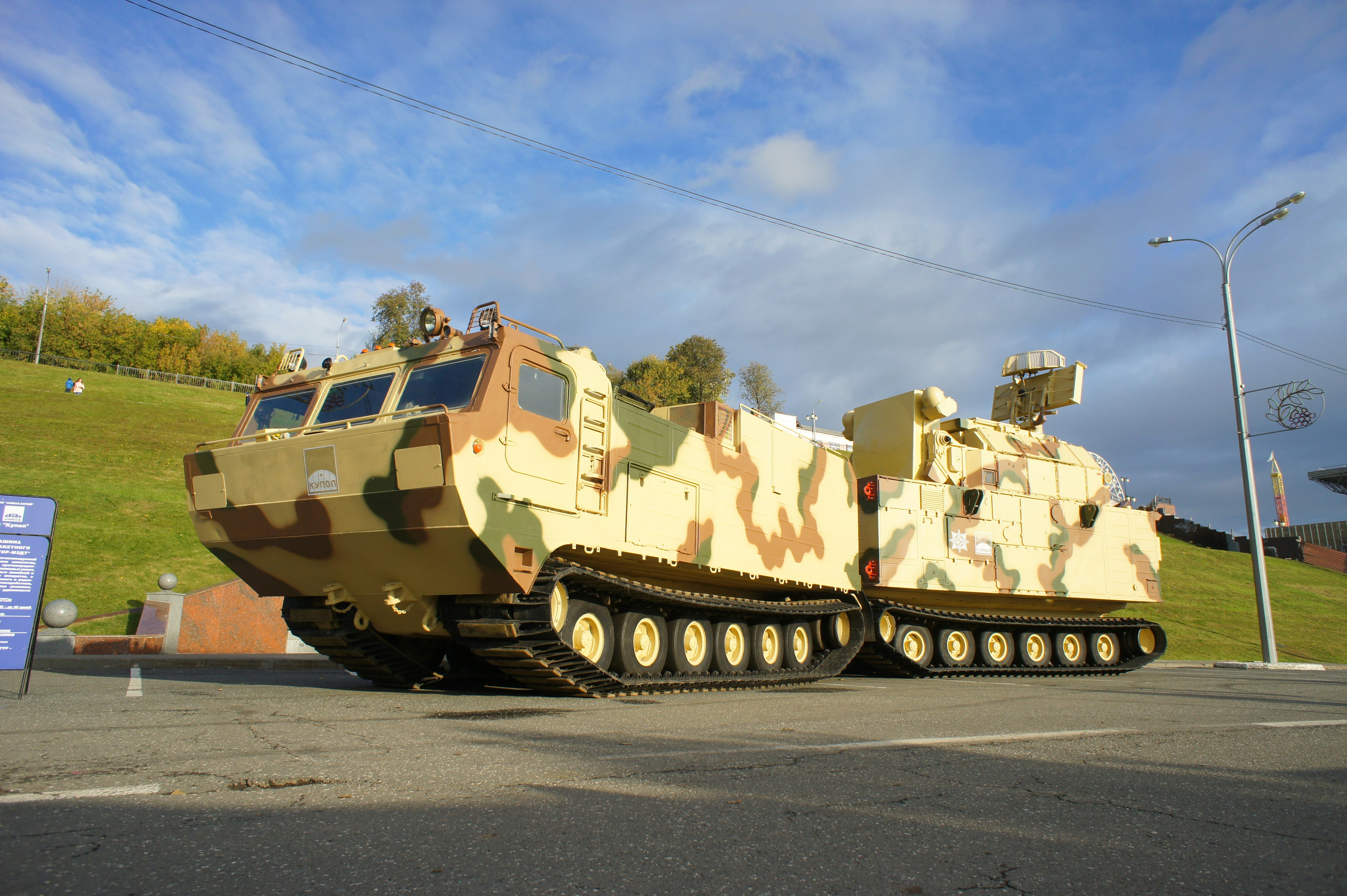A camouflaged military vehicle with tracks parked on asphalt near a grassy hill