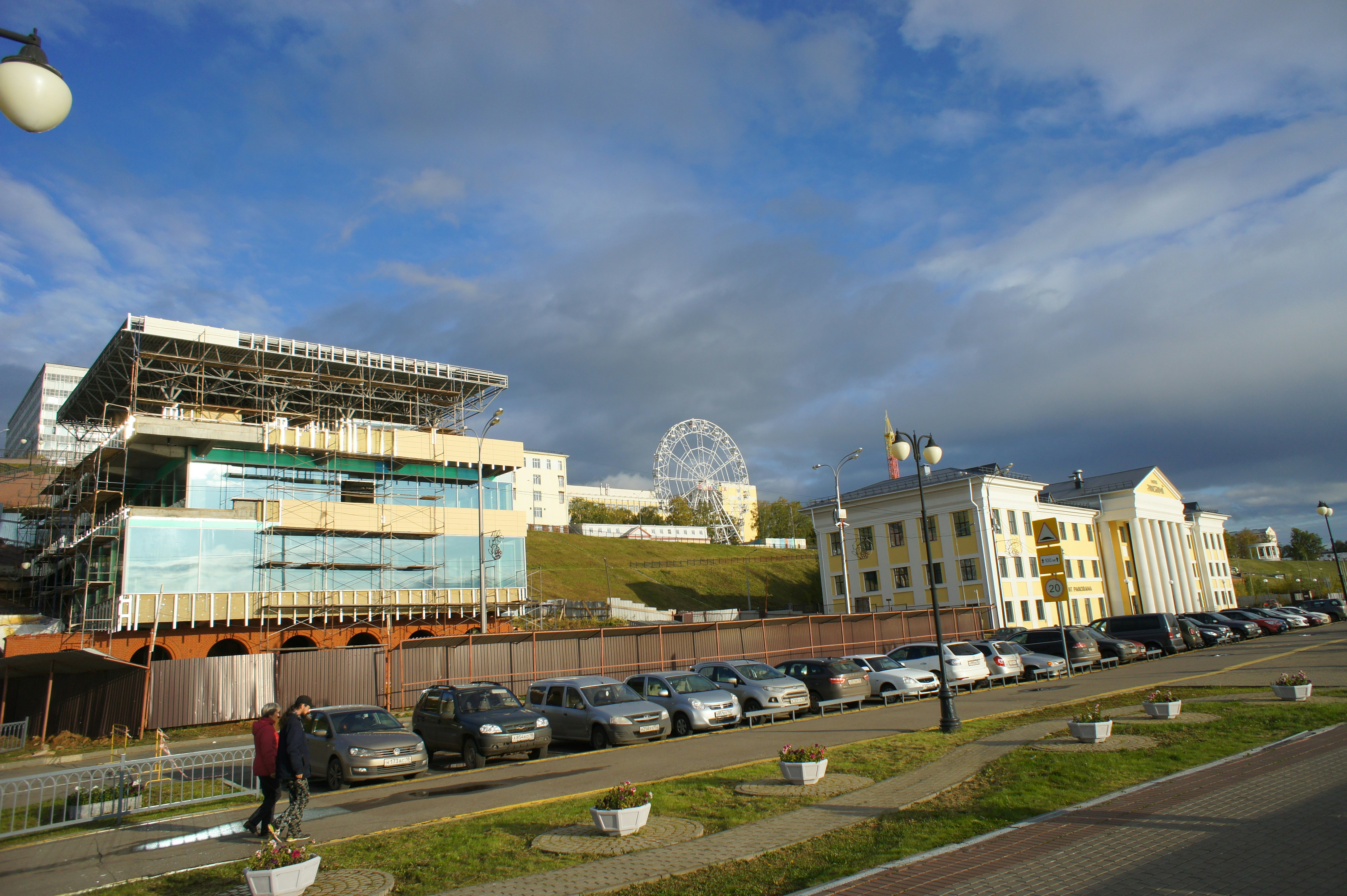 A ferris wheel behind a construction site and yellow buildings near a parking lot