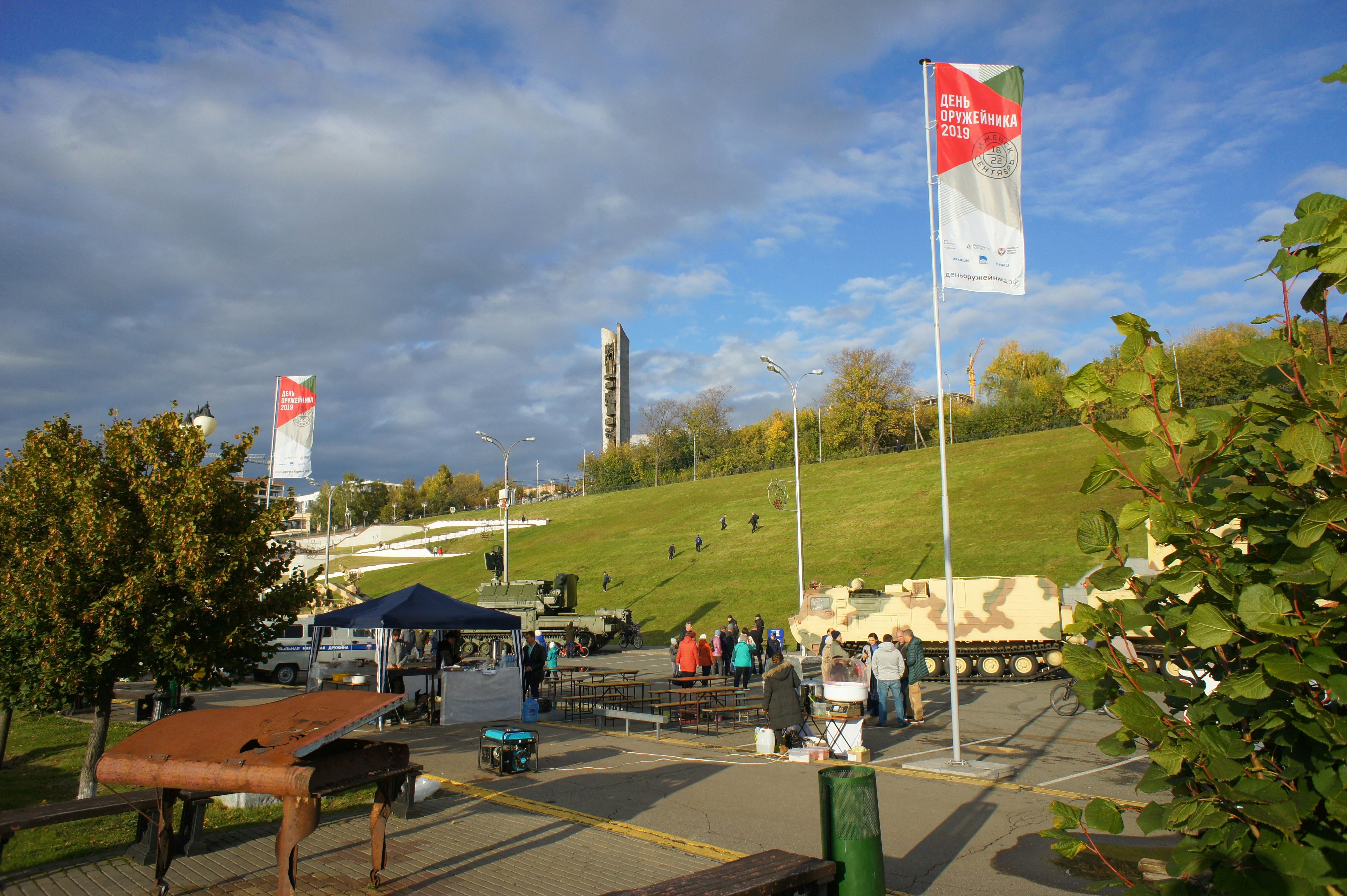 People gathered in an outdoor plaza with military vehicles and banners under a cloudy sky