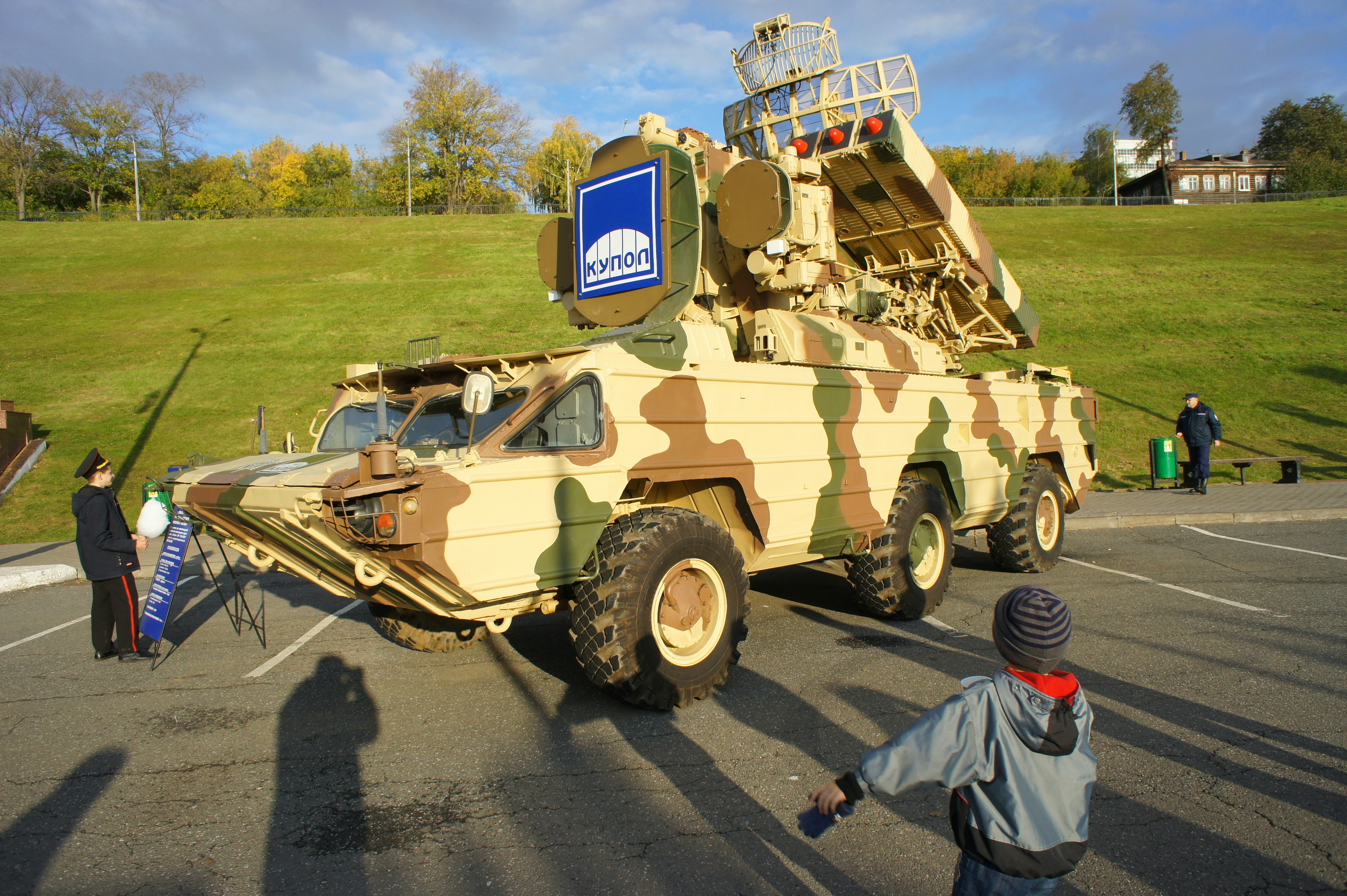 A child looking at a camouflaged military vehicle parked on an asphalt lot