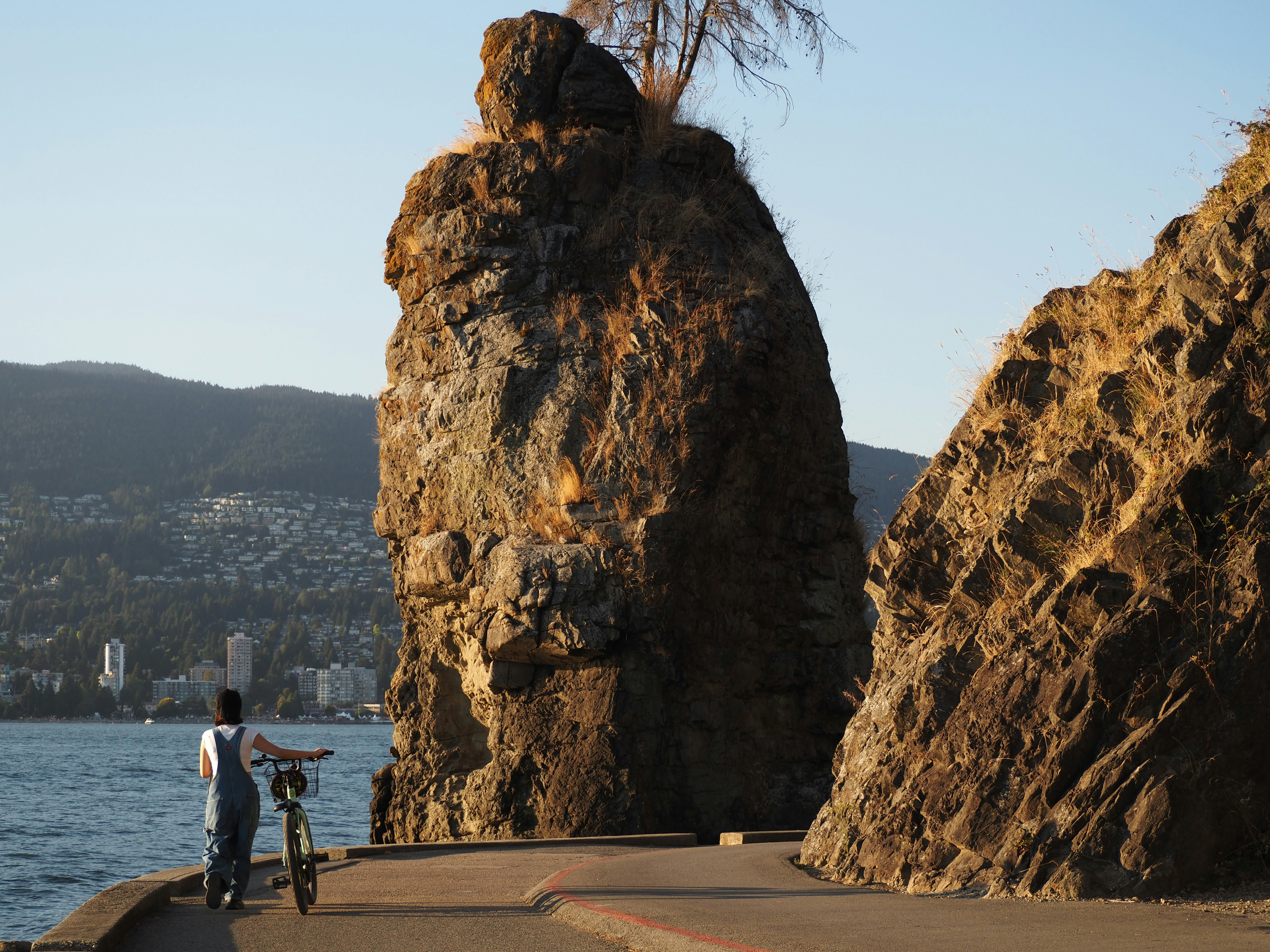 A rider walks a bike along the Stanley Park Seawall past the sea stack off the park's west shore in low evening sun.